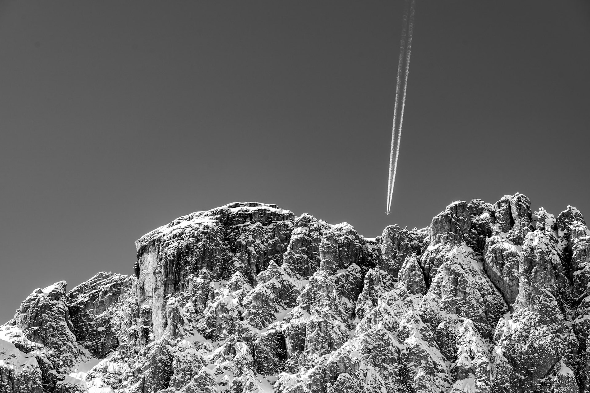 A condensation trail bisects a clear winter sky above the jagged, snow-dusted limestone pinnacles of an alpine ridge. The diagonal of the contrail introduces a human scale and quiet tension against the raw geological mass below. Black-and-white rendering strips the scene to pure form and tonal contrast.