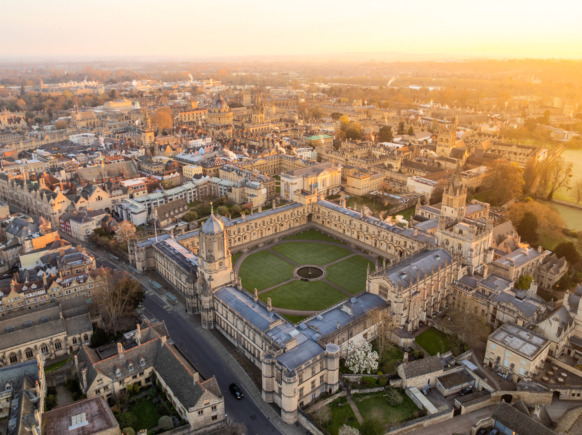 Christ Church College occupies the centre frame at low sun, its Tom Quad lawn and fountain rendered crisp from altitude. The warm raking light of a March late afternoon catches the Cotswold limestone across the Oxford skyline, dissolving into haze toward the horizon. A single flowering tree at the south range adds the only botanical note to an otherwise architectural study.