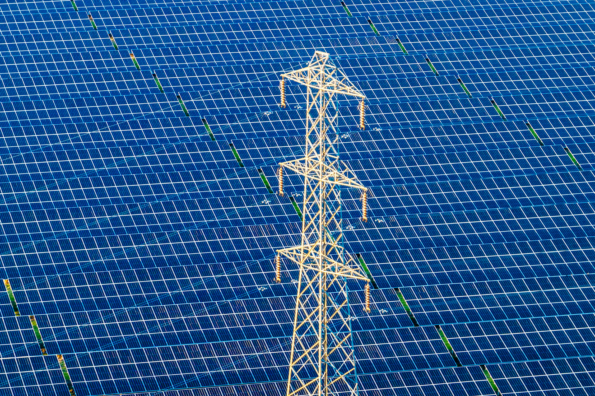 An electricity pylon stands within a large solar farm, photographed from directly above near Oxford. The aerial perspective collapses depth, reducing both structures to competing geometric patterns across a field of deep blue photovoltaic cells. The warm lattice of the steel tower reads as an anomaly against the relentless regularity of the panels beneath it.