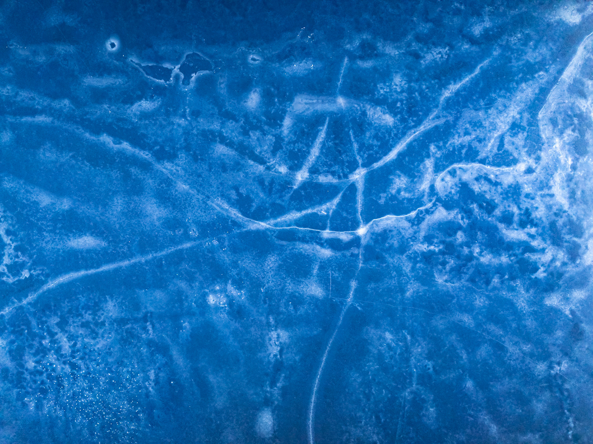 A network of stress fractures radiates across the surface of frozen ground near Nethy Bridge, Cairngorms, captured from directly above in late December. The monochromatic blue palette — deepened by low winter light and the drone's angle of incidence — renders the ice as something closer to celestial cartography than familiar landscape. Frost crystals and air pockets beneath the surface add granular texture at the margins.
