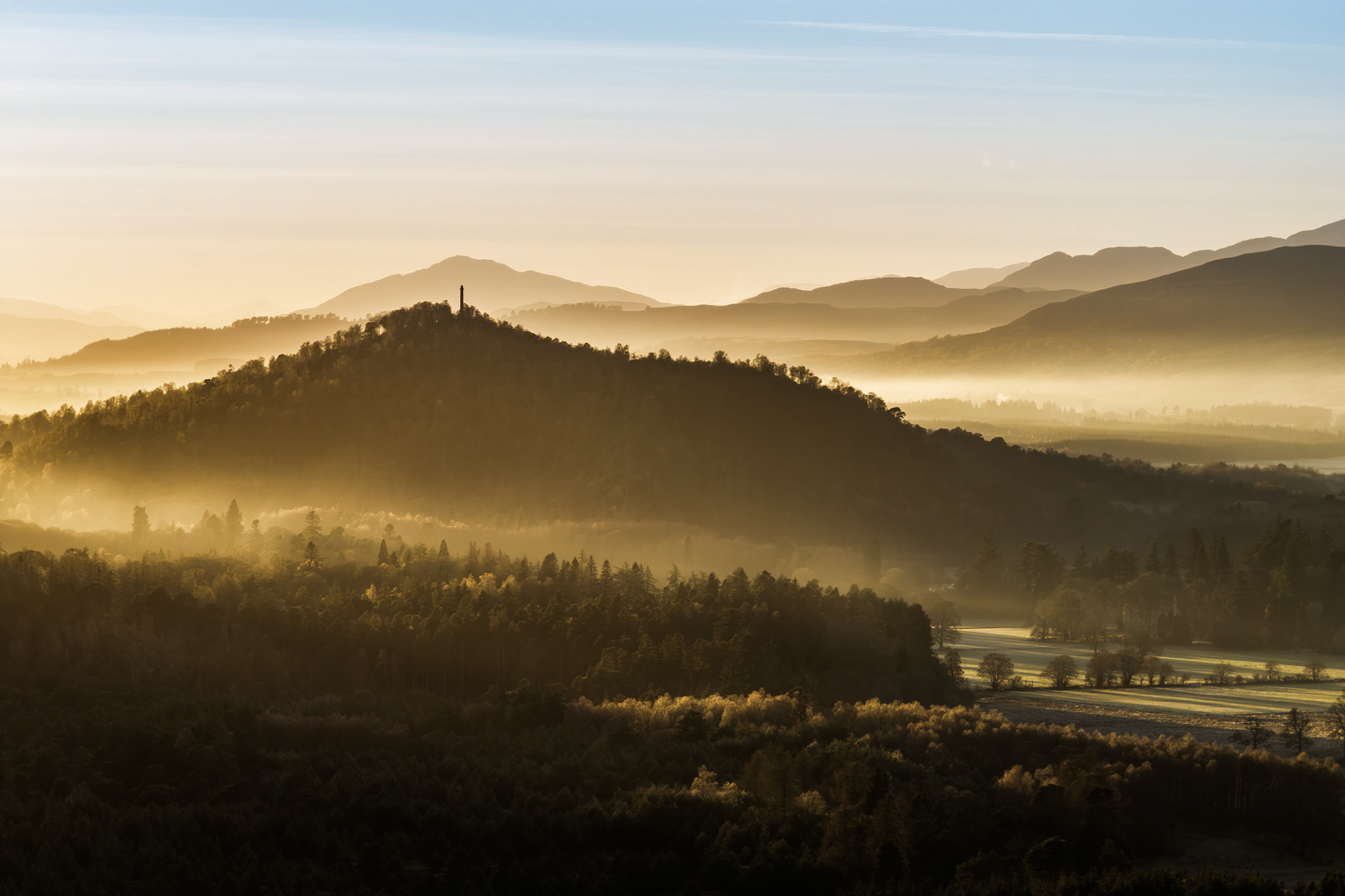 A temperature inversion pools valley mist between forested ridges near Aviemore, the Cairngorms, on a December morning. A solitary monument crowns the nearest wooded hill, its silhouette the only vertical interruption in a composition built entirely from receding planes of light and atmosphere. The distant Highland peaks dissolve into a warm, pale sky, compressing depth into something almost painterly.