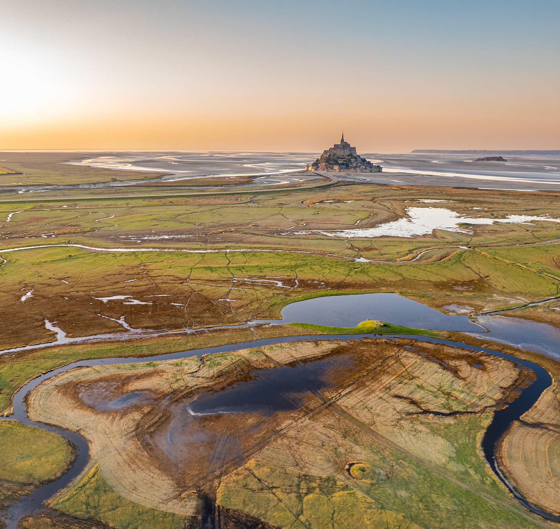 Aerial view across the salt marshes of the Baie du Mont-Saint-Michel at low tide, Normandy, France. Meandering tidal channels and waterlogged grazing land lead the eye toward Mont Saint-Michel, its abbey spire rising from the exposed bay floor at golden hour. The shallow perspective flattens the vast intertidal plain into a pattern of sinuous curves, still water, and eroded sediment.