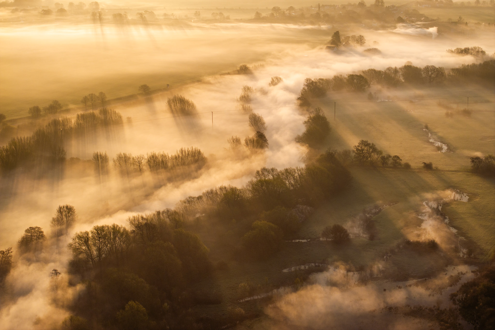 Ground fog pools along the Thames floodplain near Wallingford, pooling between lines of riparian woodland as low March sun casts raking crepuscular rays across the inversion layer. Shot from altitude at the equinox, the image resolves the valley into interlocking bands of light, mist, and bare canopy — the pastoral geometry momentarily obscured and revealed in the same breath.