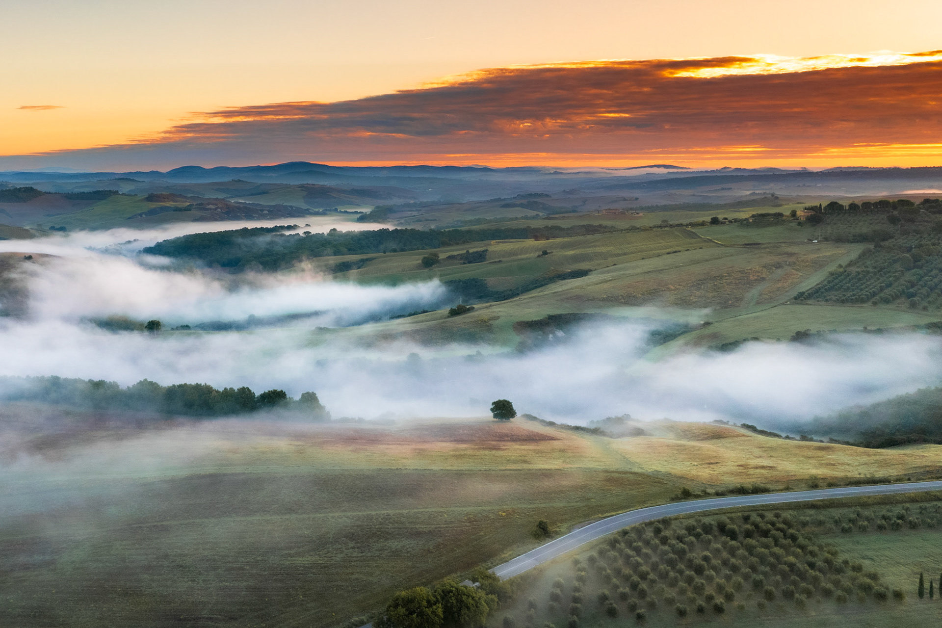 Ground-level inversions pool between the rolling hills of the Val d'Orcia at dawn, the mist threading between tree lines and farmland as the sky ignites above the distant Apennine ridge. Shot from altitude, the layered recession of fog, hill and cloud gives the scene its depth. Autumn colour is still emerging in the canopy below.