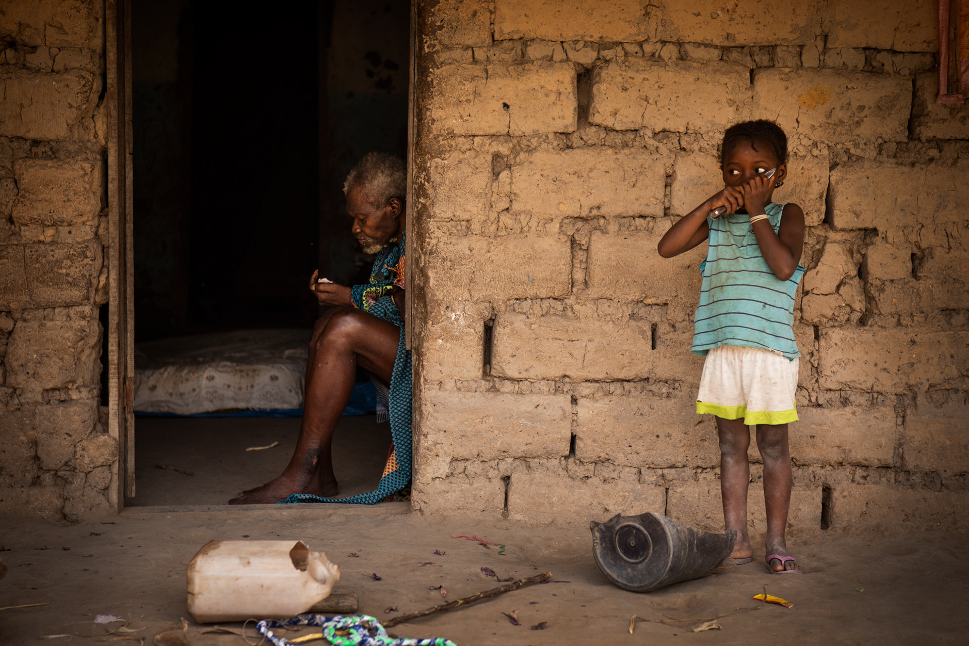 bambina che gioca con un coltello accanto al nonno che mangia, in un villaggio balanta in guinea bissau, africa. Kid playomg with a knife with his grandfather eating an apple, in balanta village, guinea bissau