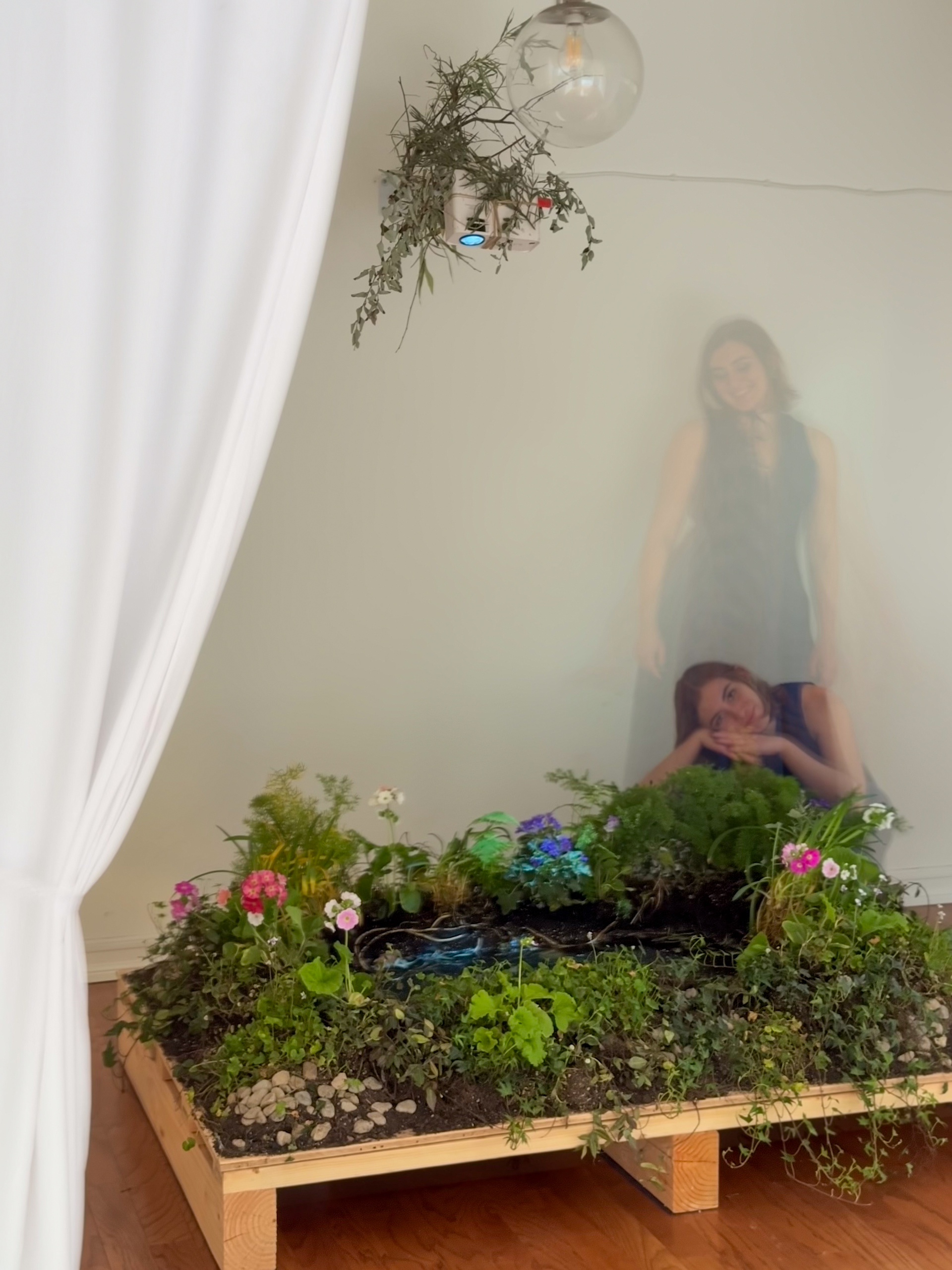 Sophia Wolfe stands behind her installation, "Icolette Pond", an indoor pond with blooming pink and purple flowers sitting upon a wooden platform. A projector adorned with leaves and tree branches is above, facing down, projecting an image into the water of the pond. The water glows blue. A white curtain is brushed to the side on the left half of the image. The image is double exposed, Sophia is both standing up and on the floor resting her head upon the flowers. This photo was taken at Alt Projects for the exhibition "Staring Contest". The photo was taken by Daniel Bilezikian.