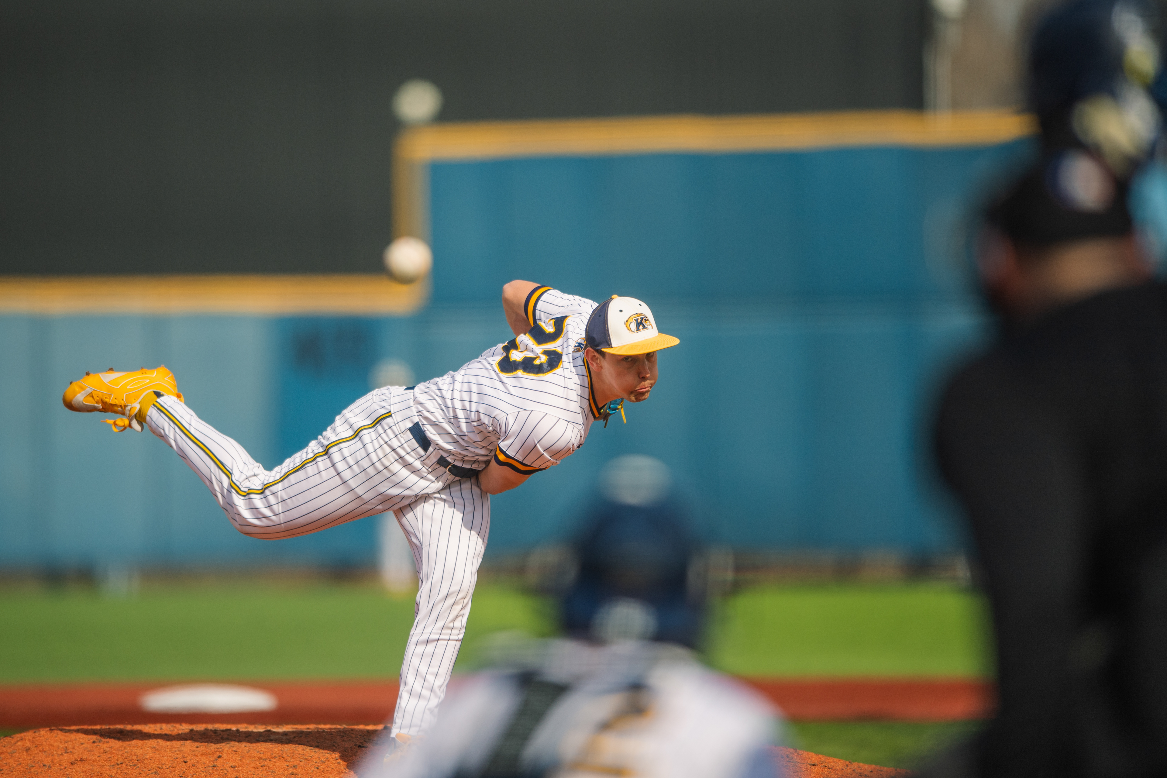 Kent State's #23 fires through his delivery against Rhode Island, the ball trailing through the upper frame as the umpire sets behind the plate.