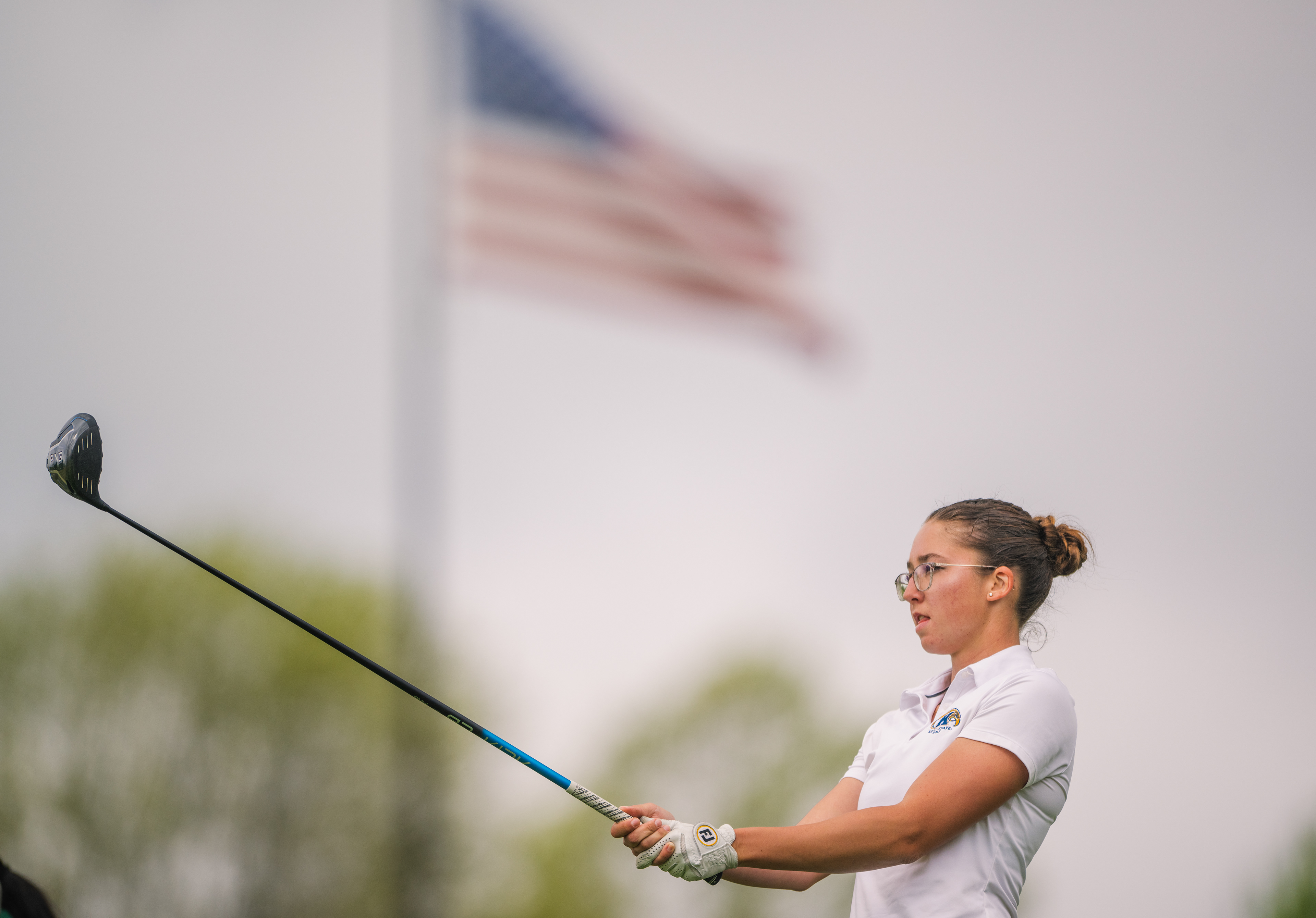 A Kent State golfer eyes the fairway with a driver at rest. The American flag blurred overhead in the hazy afternoon sky, framing the moment before she steps to the tee at the MAC Championship.