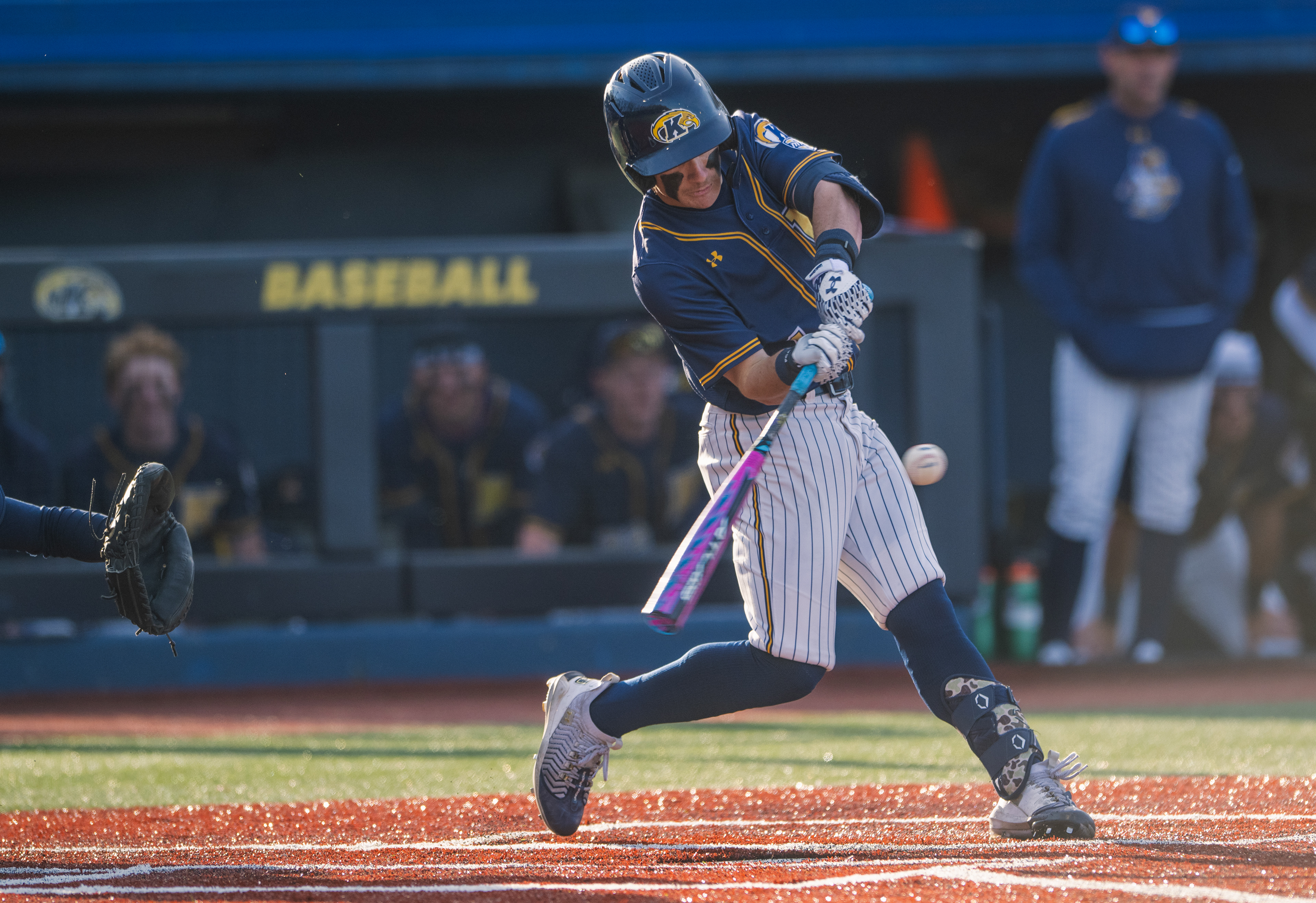 Ball meets bat as a Kent State hitter makes contact in front of the home dugout, the ball still visible off the barrel as the catcher braces for the result.