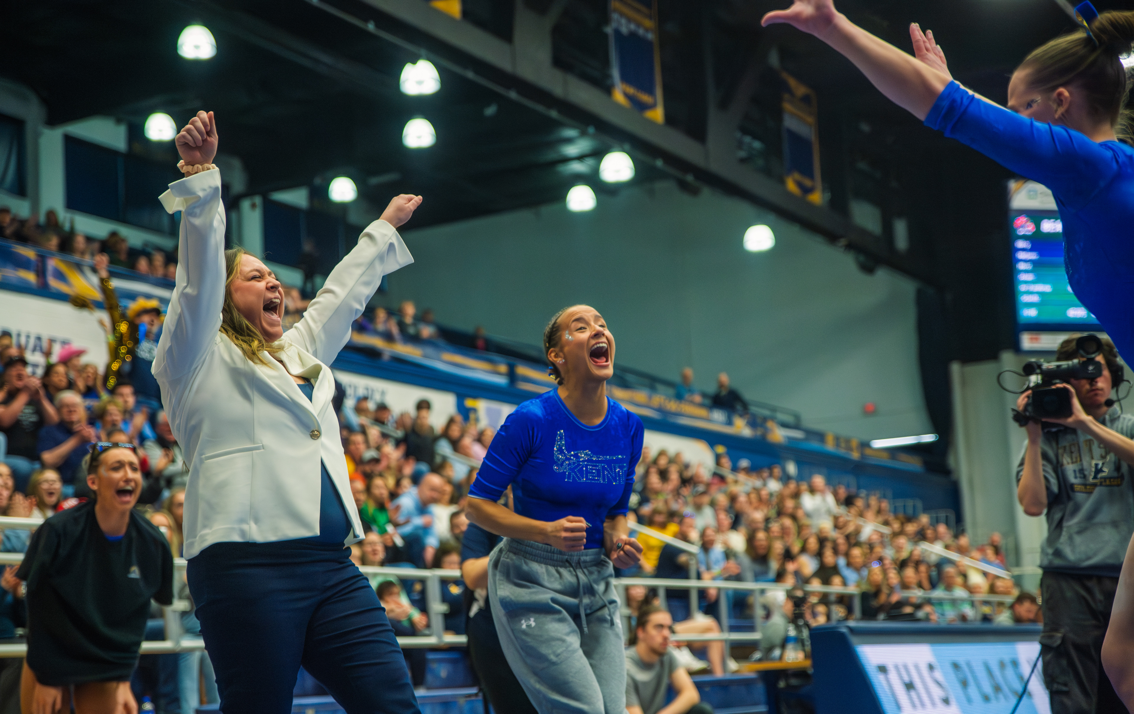 A coach and gymnast scream in unison as a teammate sticks her landing. The packed MACC crowd rising behind them, the scoreboard and camera crew framing a meet at full intensity.