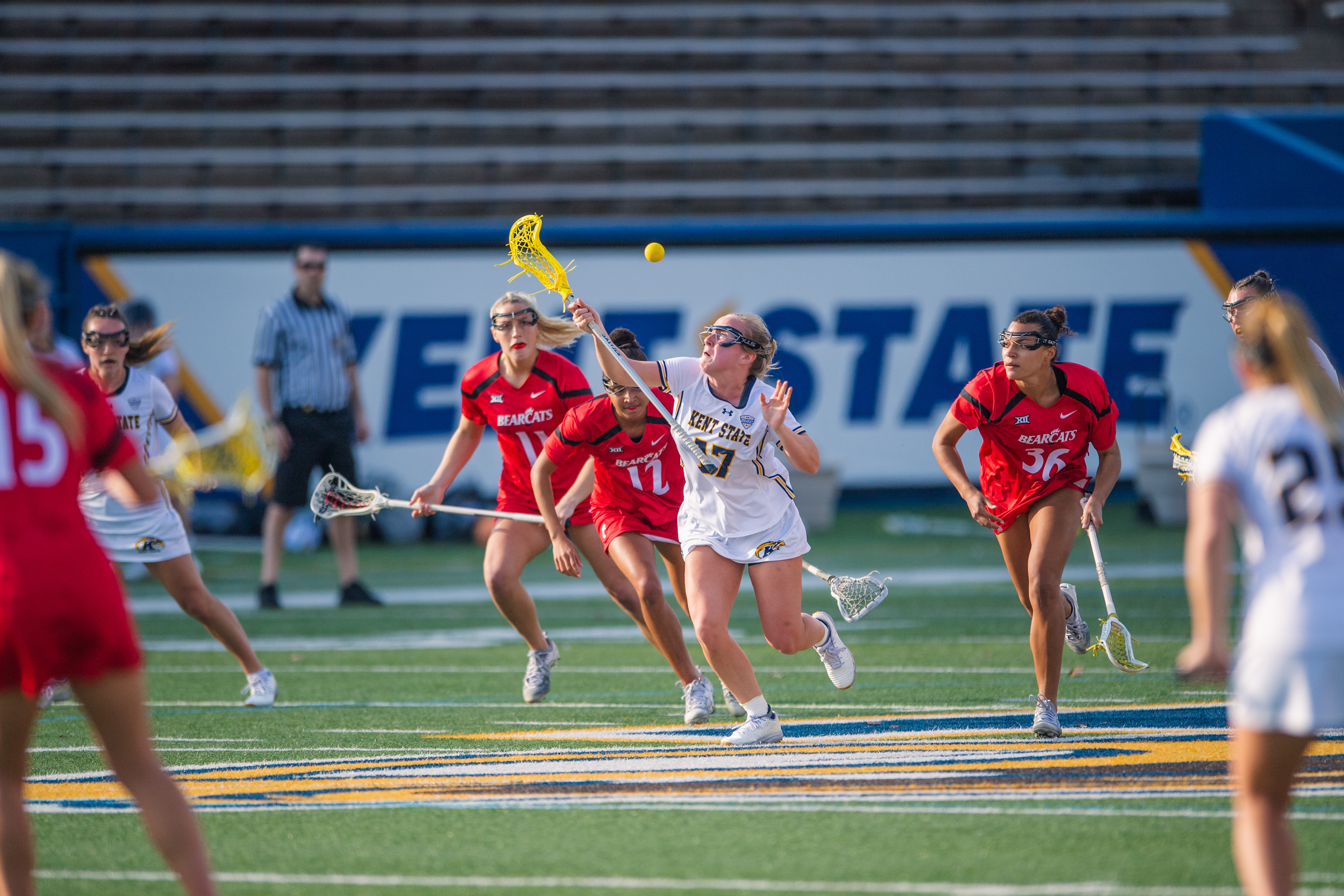 Kent State's #27 stretches to corral a loose ball with Cincinnati closing from both sides with the yellow lacrosse ball visible mid-air above the chaos of the contested possession.