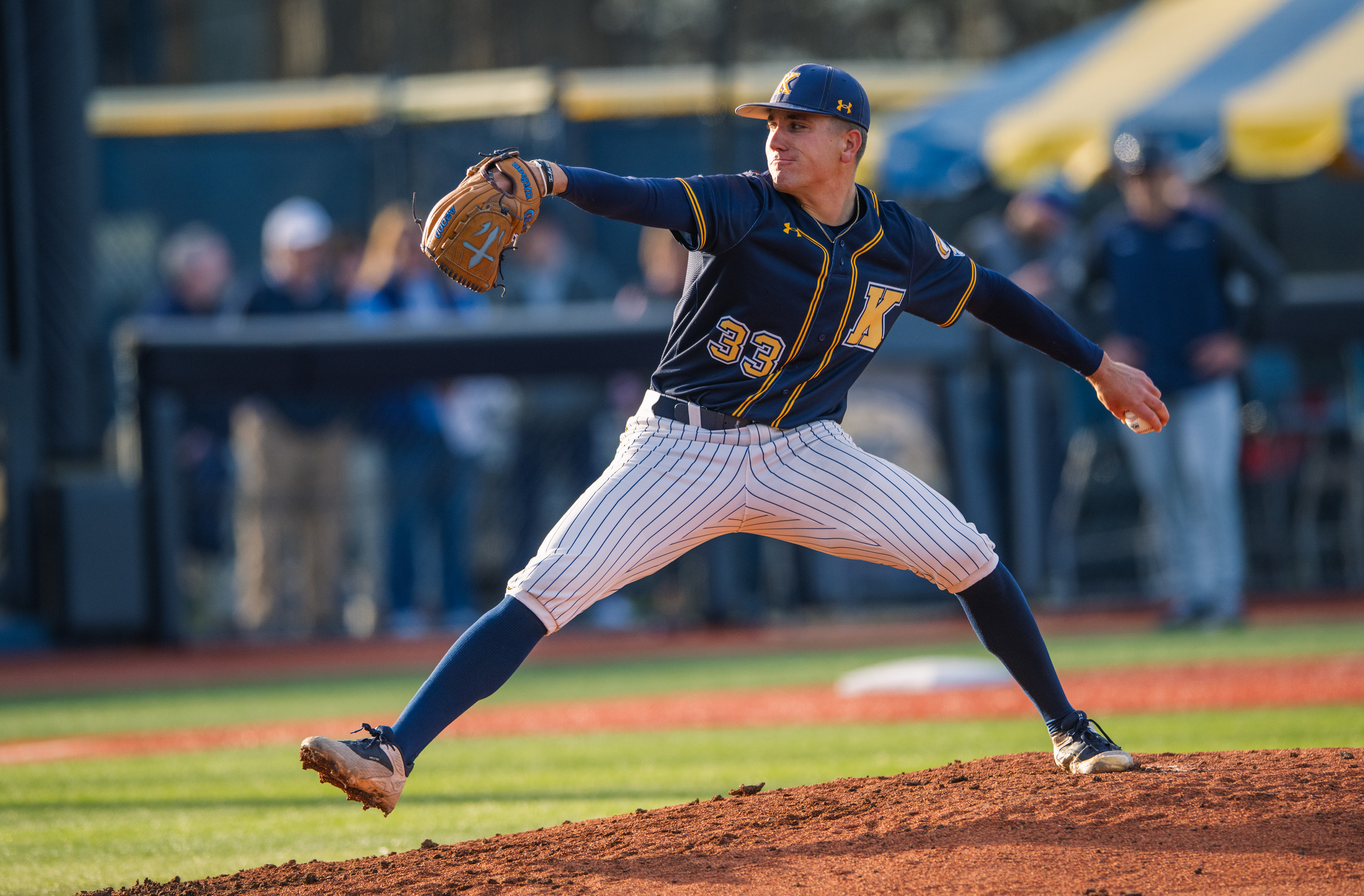 Kent State's #33 extends fully through his delivery in the long afternoon light. The golden hour catching the pinstripes and the red clay in equal measure.