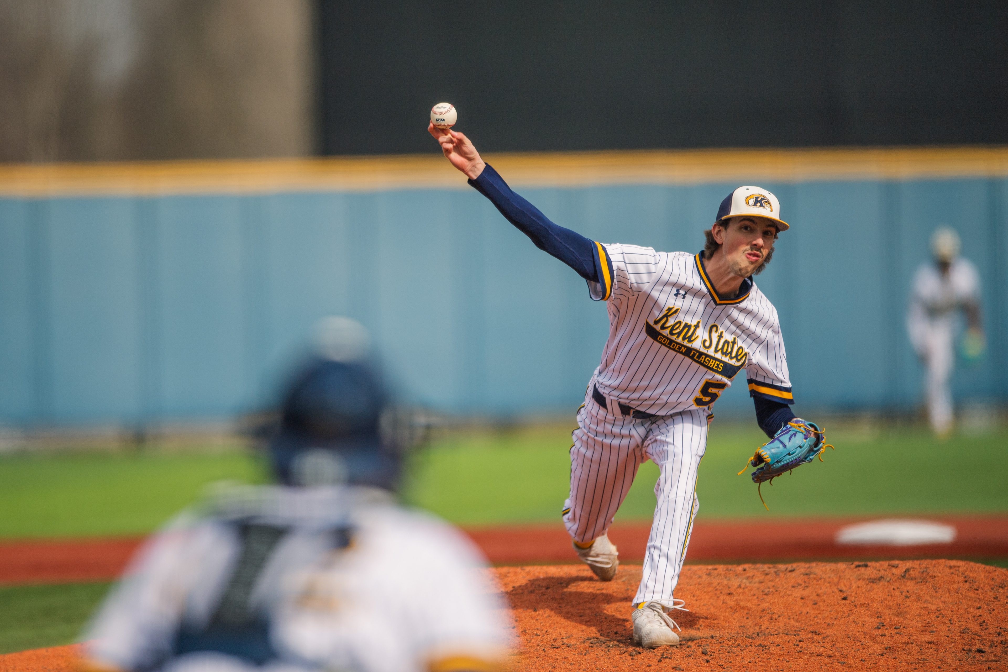 A Kent State Golden Flashes starter holds the ball at the top of his wind-up, the catcher crouched and waiting in the foreground. The tension of the moment before the pitch.