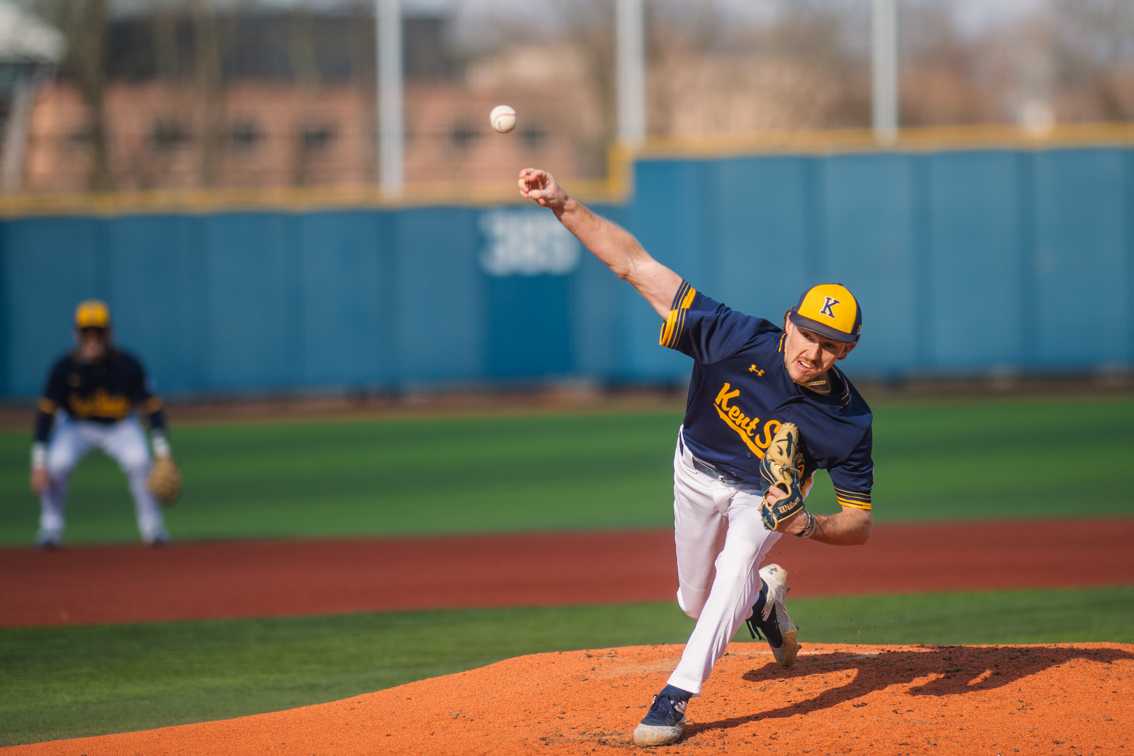 A Kent State starter fires toward the plate against Rhode Island. The ball clearly visible mid-flight as the shortstop holds his position in the background.