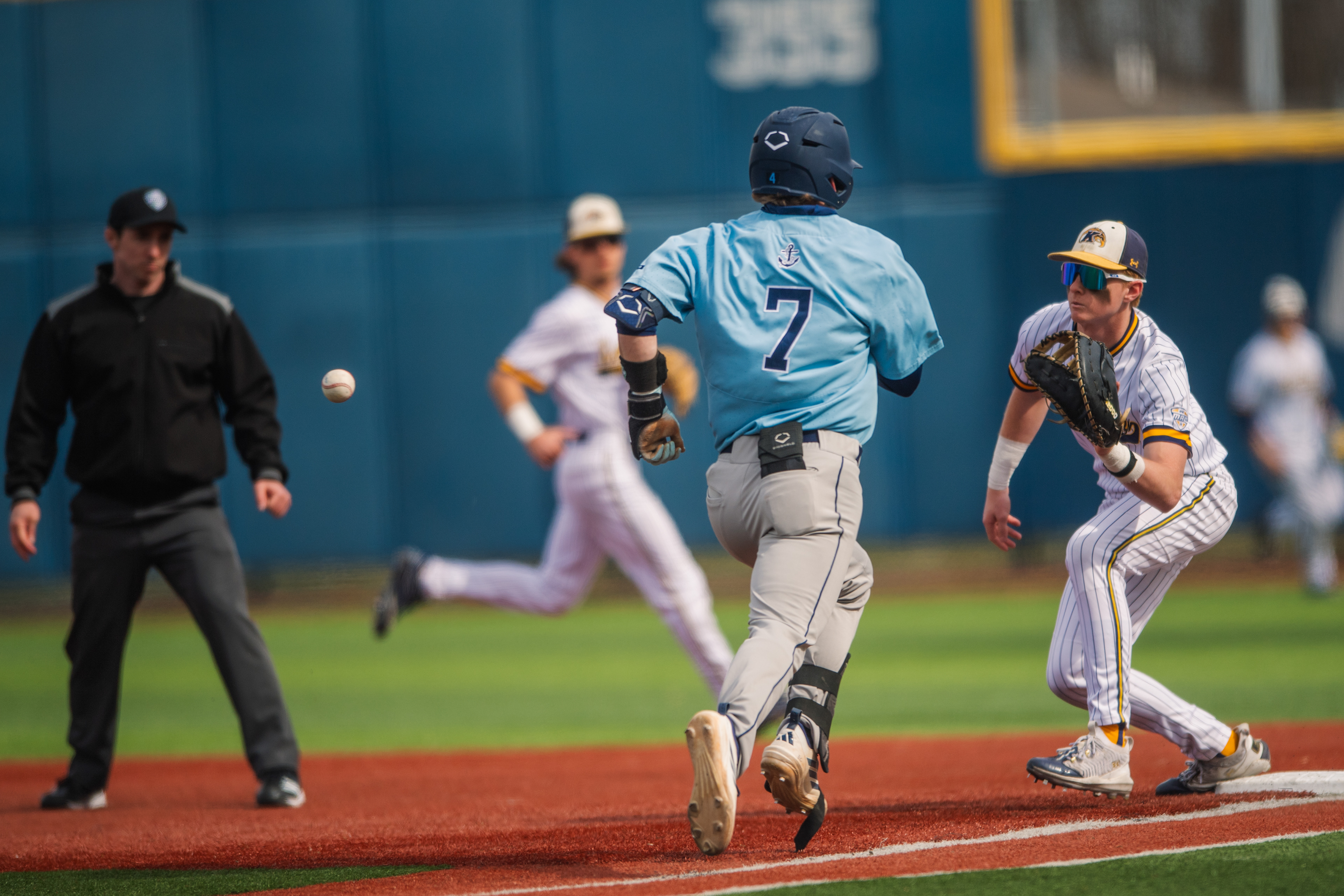 A Kent State first baseman sets to receive as a Rhode Island runner charges down the line. The ball between them, outcome still undecided, umpire leaning in to make the call.