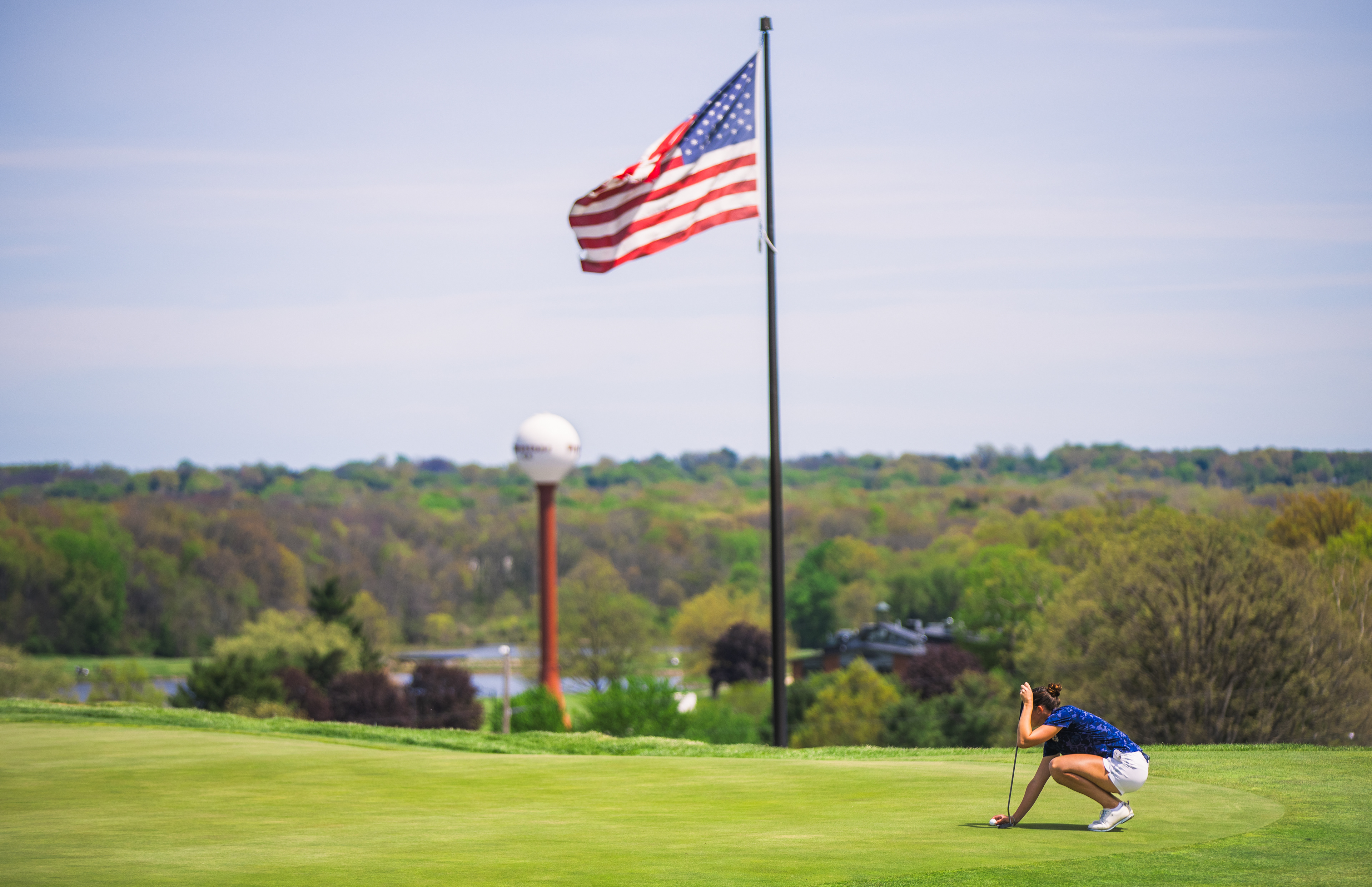 The American flag catches the wind as a Kent State golfer reads the line on a crucial green. The Ohio tree-line and water tower stretching across the horizon behind her at the MAC Championship.