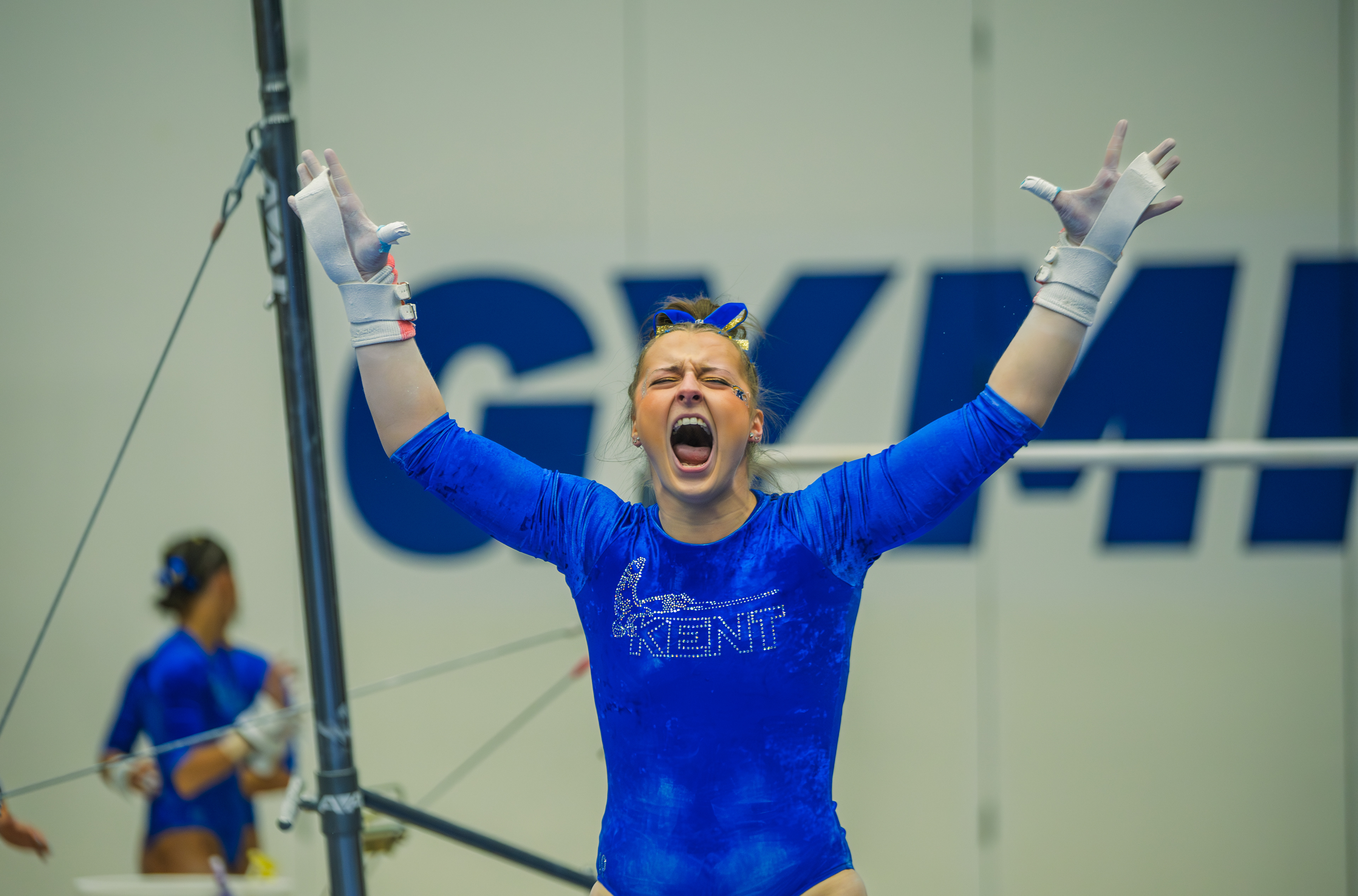 Arms raised and voice open as a Kent State gymnast erupts after sticking her bar routine, the word "GYMN" blazing across the wall behind her as the moment lands.