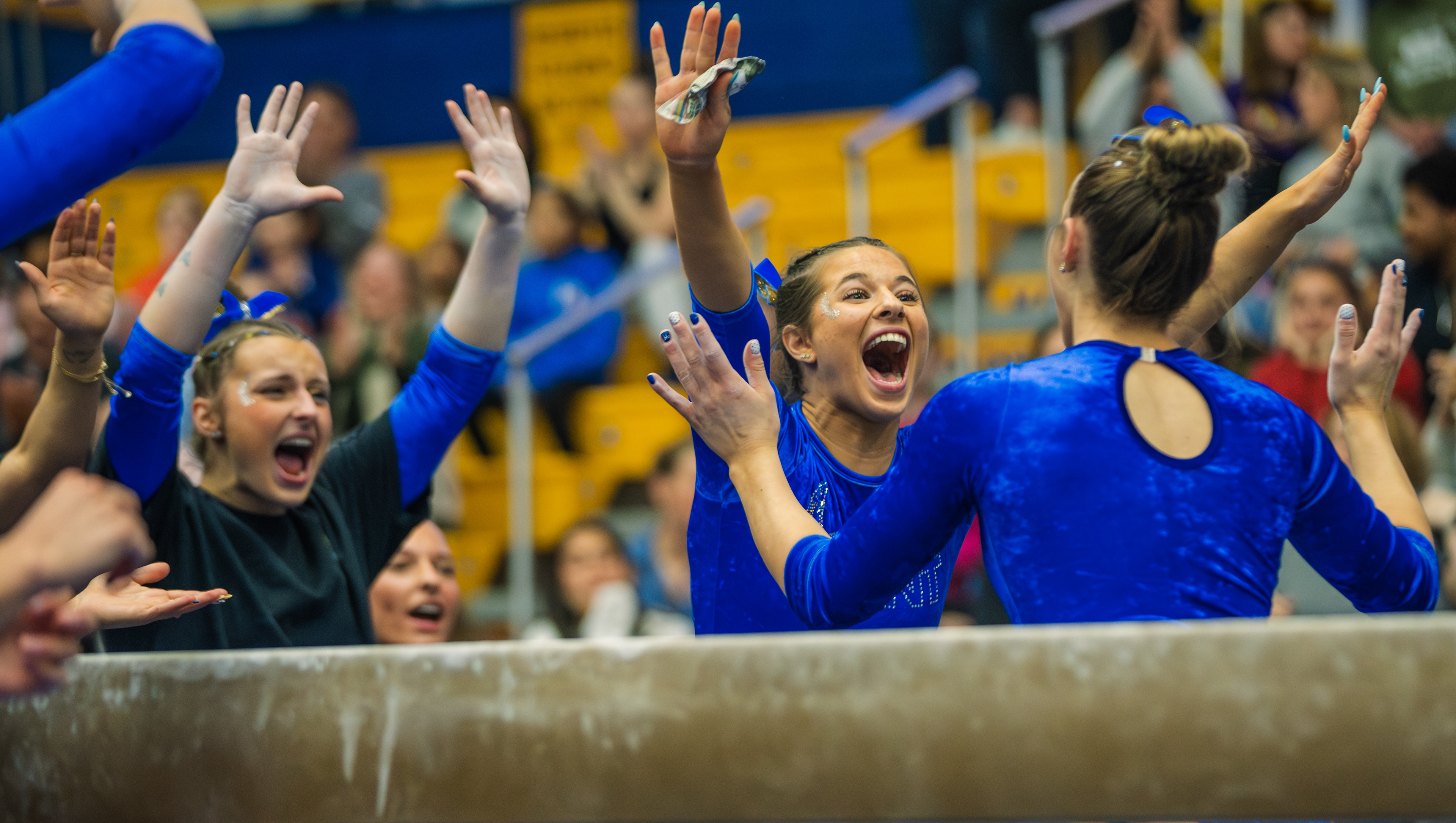 The Kent State gymnastics team swarms the beam to celebrate a teammate's score. Hands raised and mouths open as the meet swings their way in front of a packed arena.