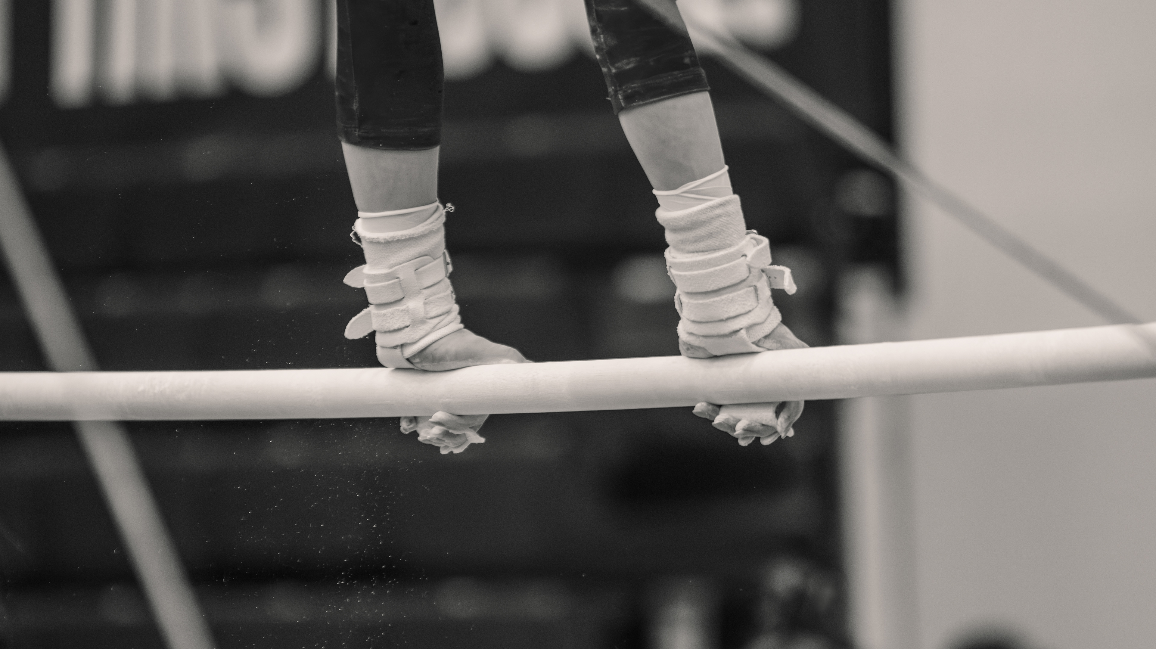 Chalk dust suspended in the air and white grips locked around the bar. The entire demand of uneven bars compressed into a single detail, nothing else needed.