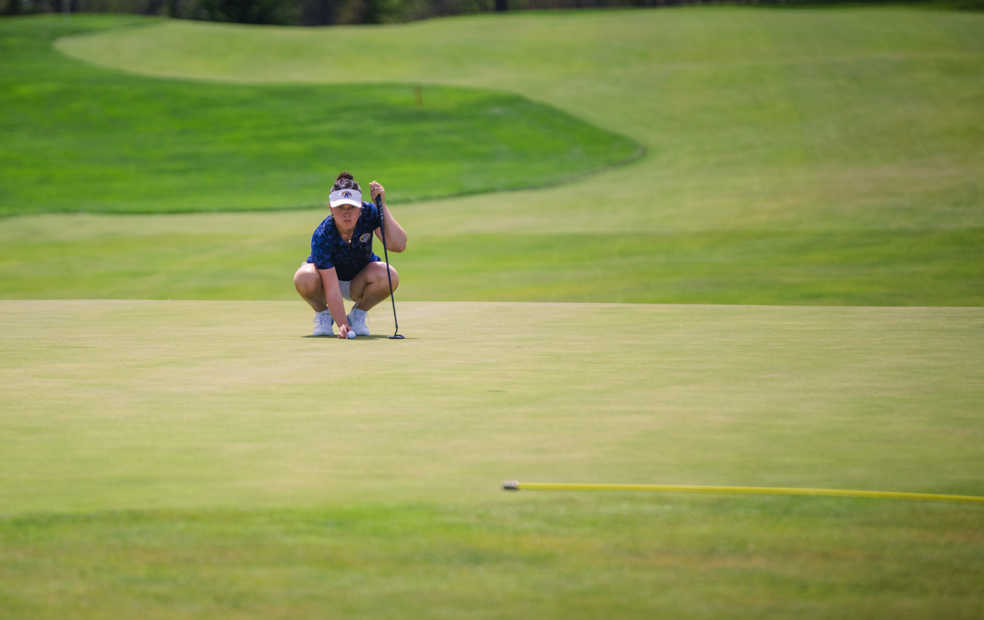 A Kent State golfer surveys a lengthy putt at the MAC Championship. The pin and yellow boundary rope anchoring the foreground as the fairway sweeps behind her into open space.