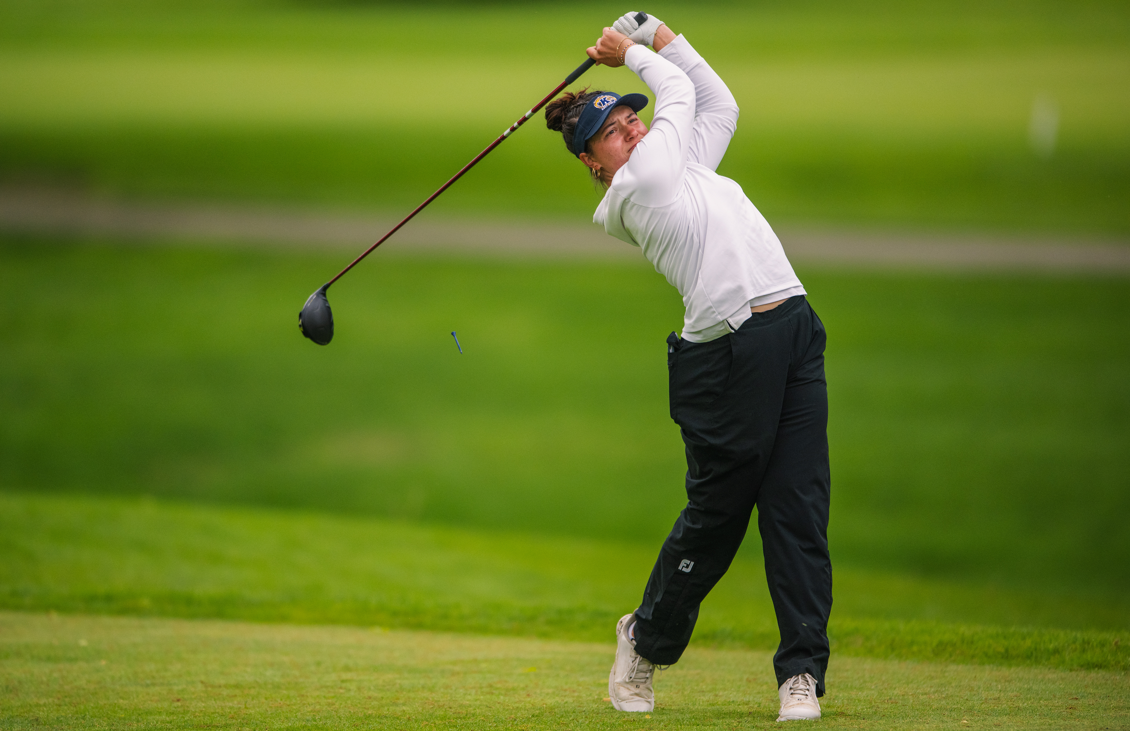 A Kent State golfer uncoils into a full driver follow-through, the tee launching skyward as she tracks the flight of her drive. Clean green bokeh framing the moment at the MAC Championship.