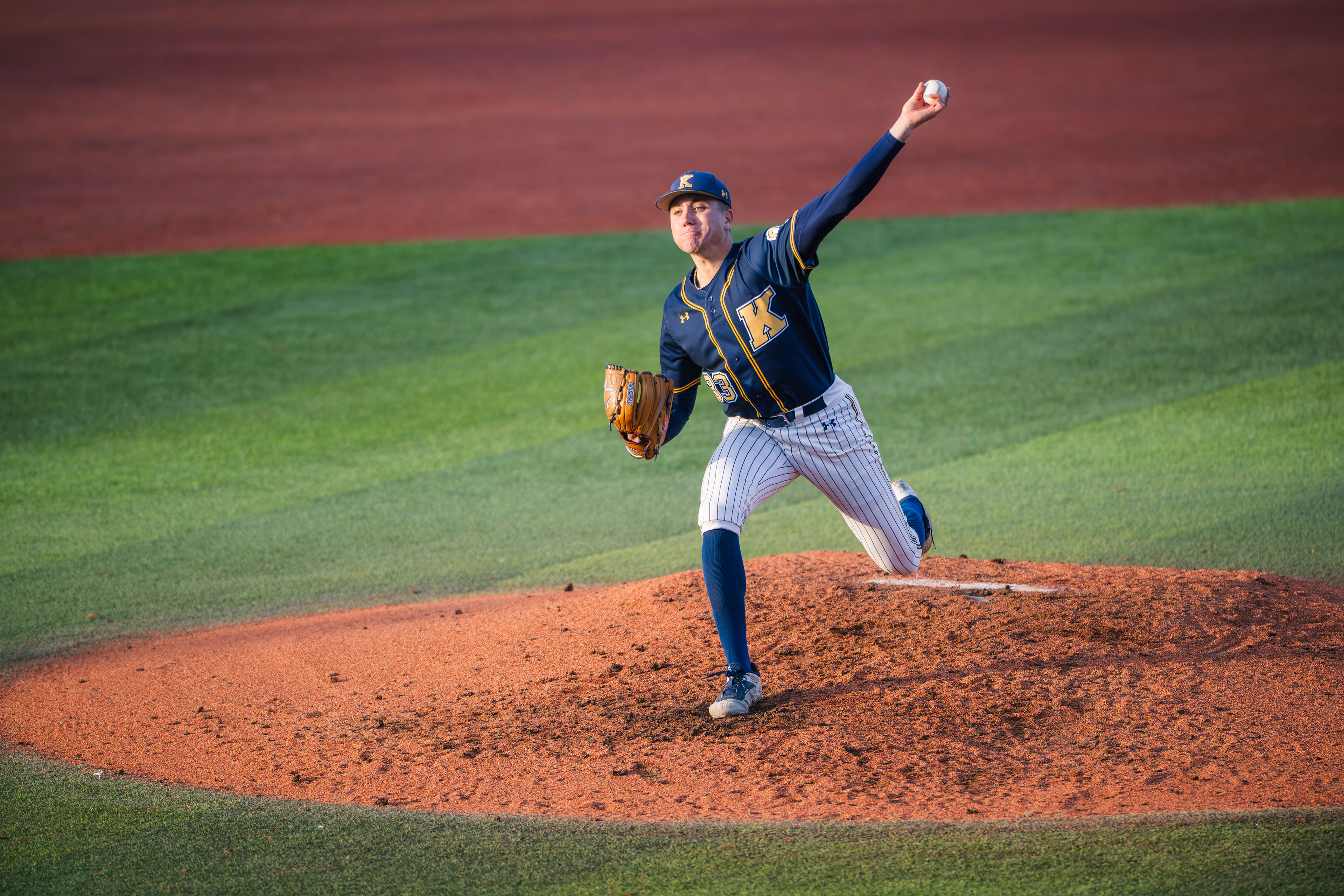 Kent State's #33 winds into his delivery as the last of the afternoon light falls across the mound. The red clay, green grass, and pinstripes in perfect alignment.