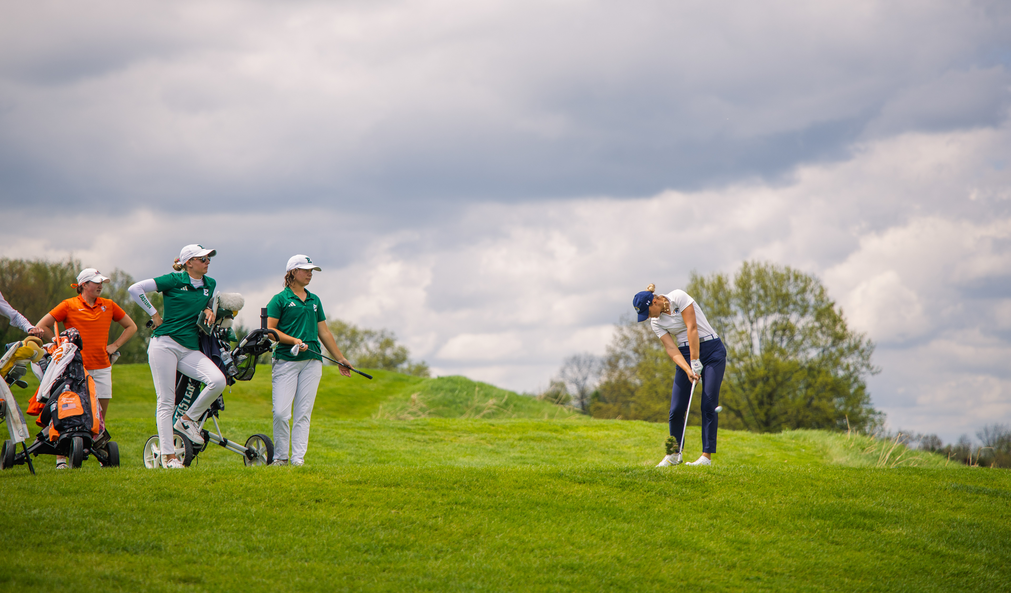 Under a building storm sky, a Kent State golfer works a chip from a hillside lie while the Eastern Michigan contingent watches on. The MAC Championship field gathered in tense silence.