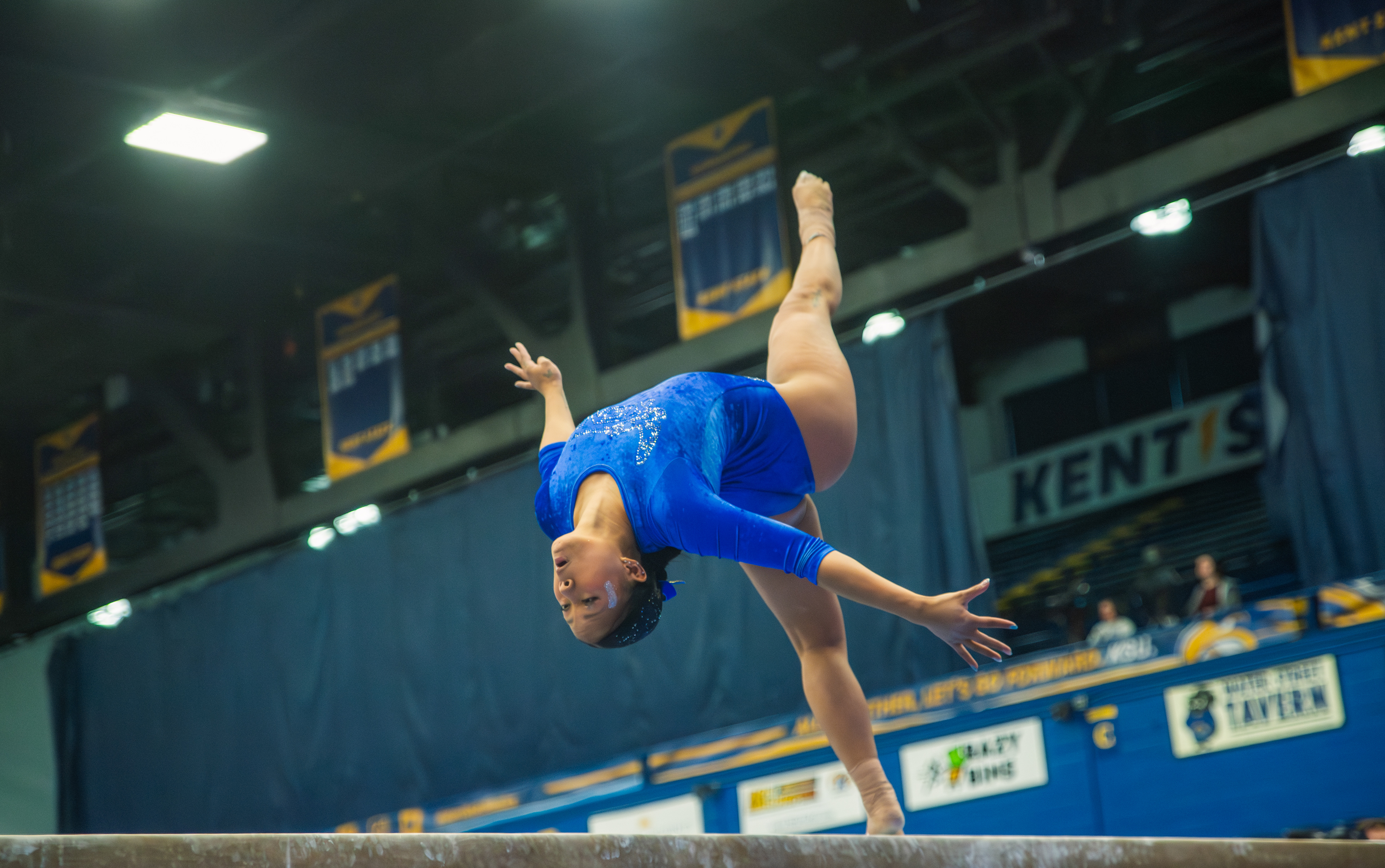 Inverted and fully committed. A Kent State gymnast works through a back walkover on the beam, championship banners and arena lights hanging overhead as she trusts the four-inch surface beneath her.