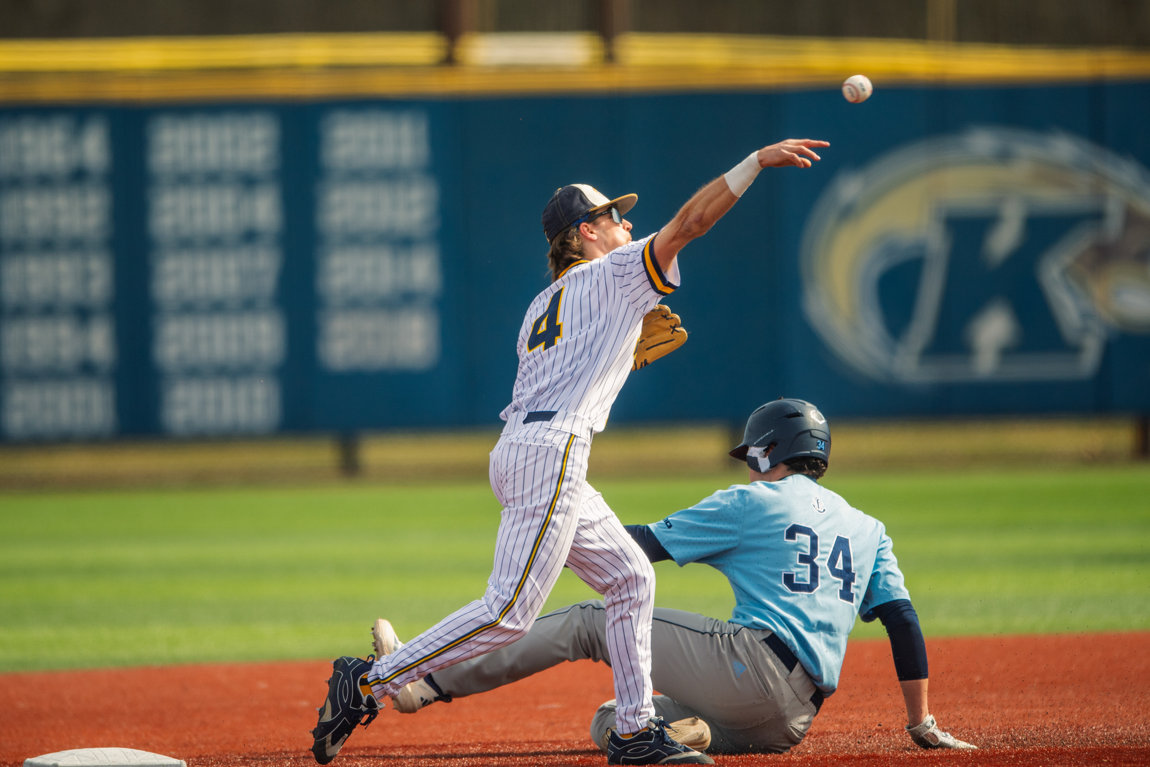 Kent State's #4 pivots at second base and fires to first with a Rhode Island runner sliding in beneath him. The ball still airborne, the double play turn caught mid-execution against the championship wall.