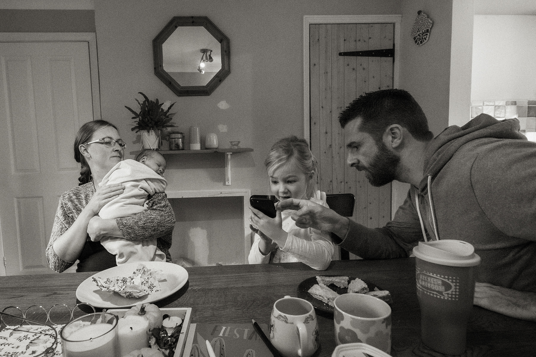 family sitting at table while father shows daughter something on mobile phone