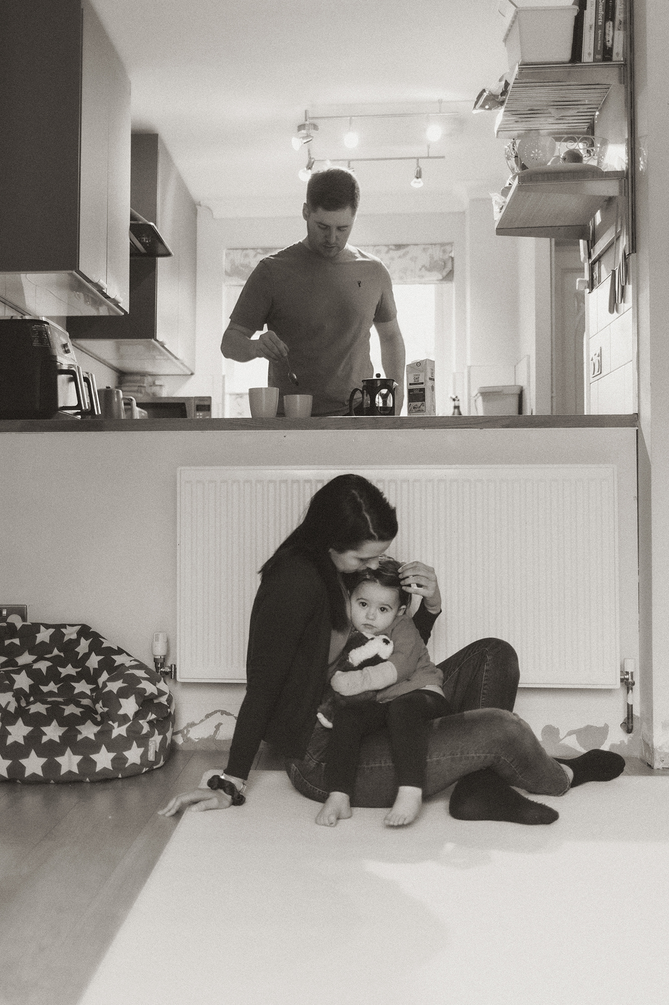 mother consoling child while father makes coffee in the background