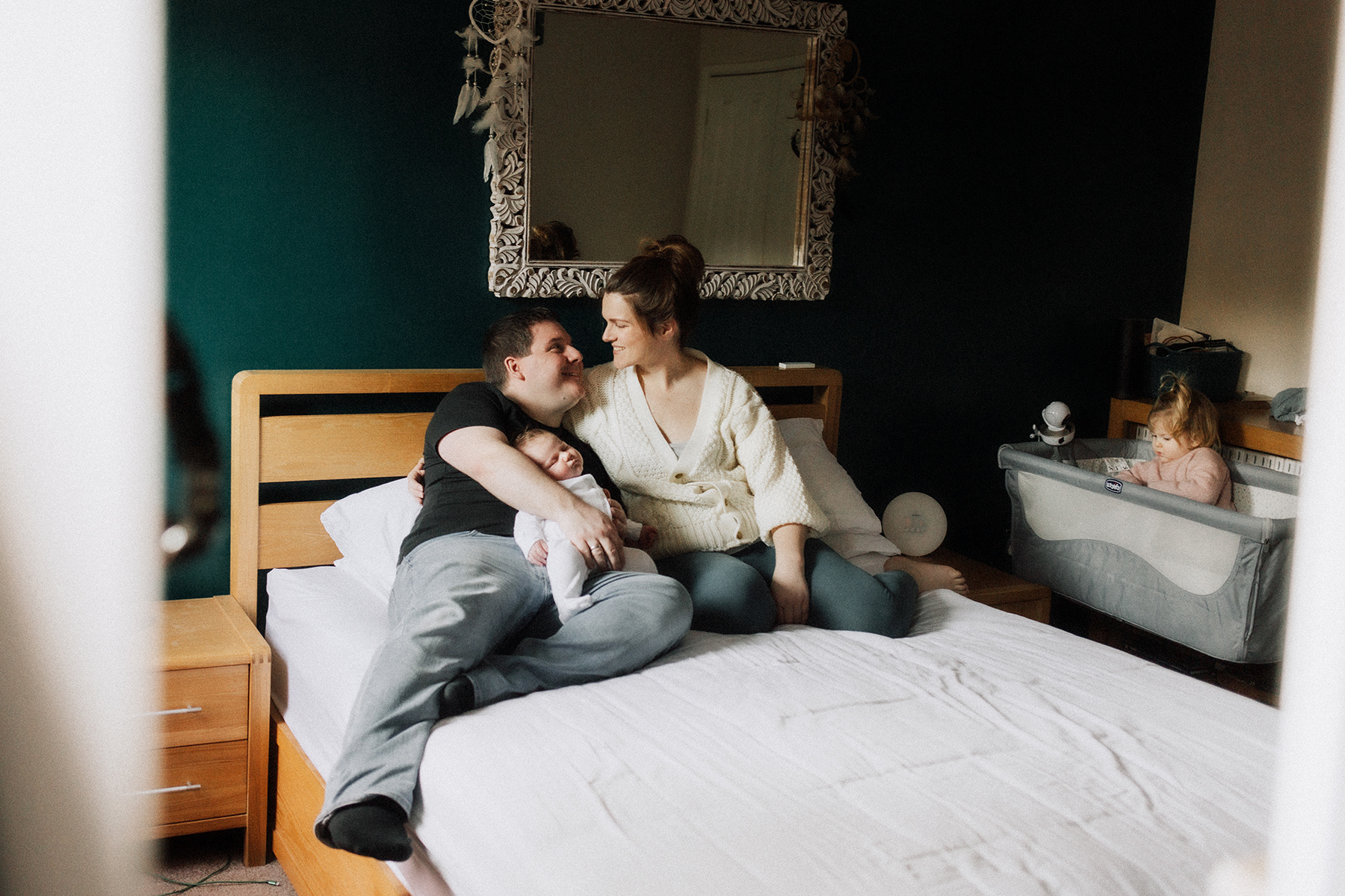 parents gazing lovingly at eachother while daughter plays in baby cot