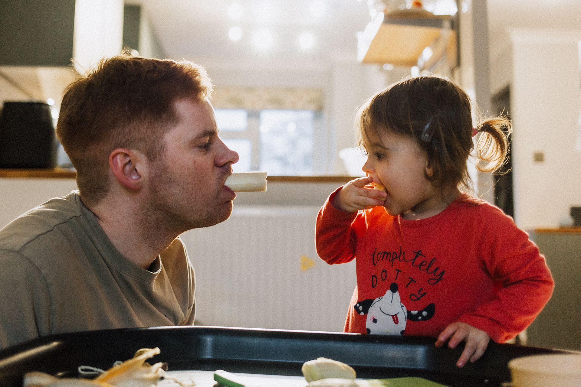 dad pulling funny face at daughter with banana in his mouth