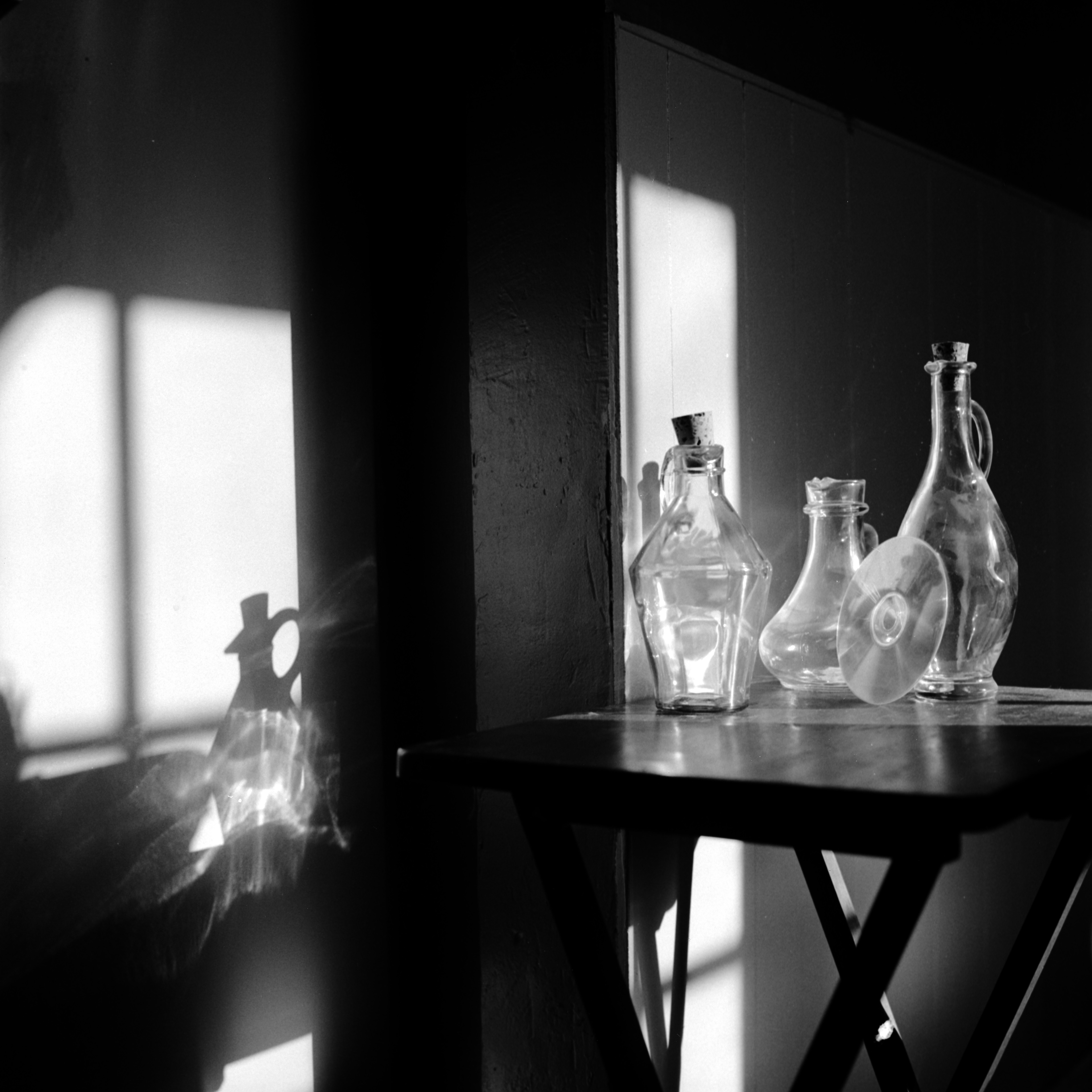A black and white image of three transparent glass bottles, two with corkscrew lids, and a CD rested in between, all of which are atop of a television tray. One of the bottles creates a noticeable shadow from being in the daylight.