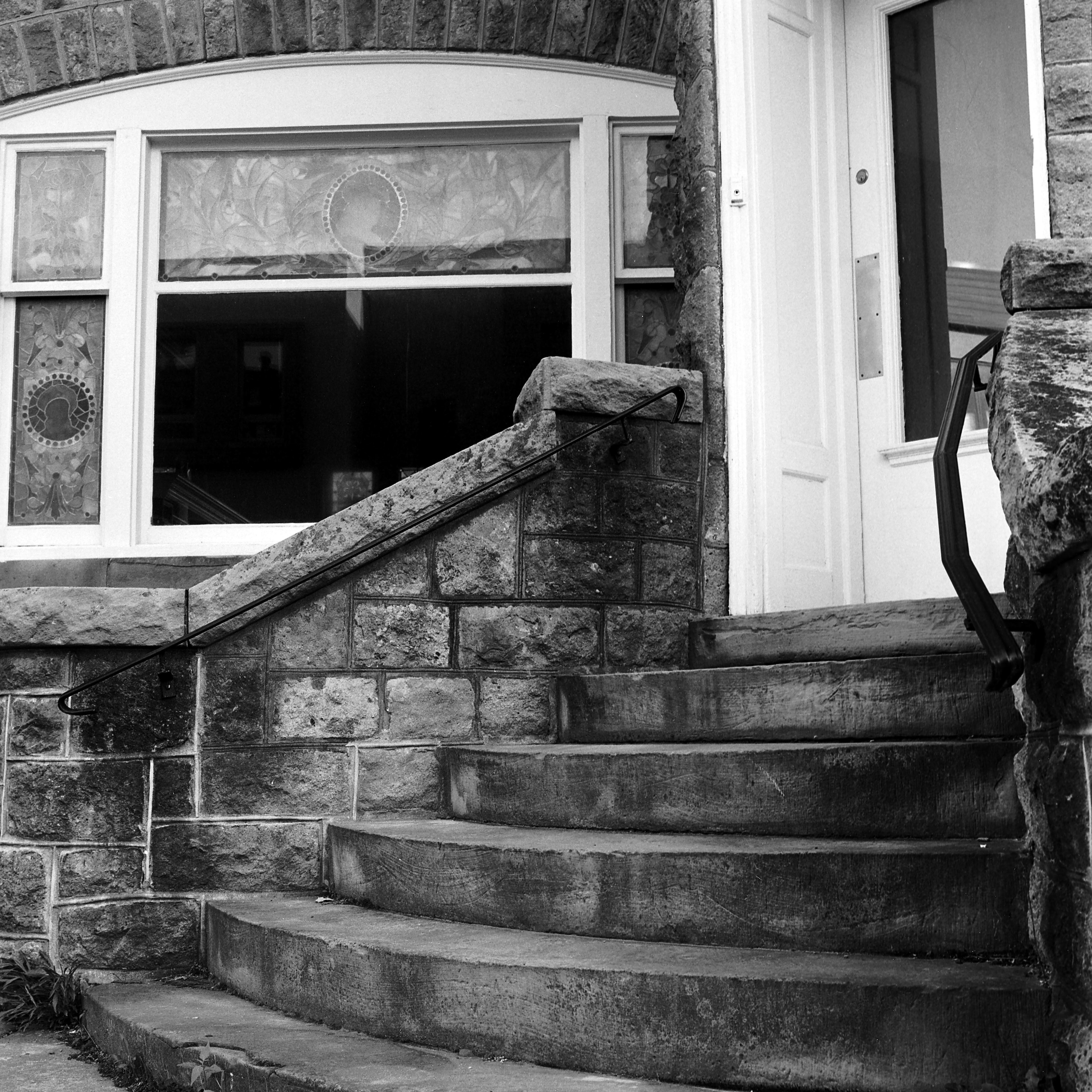Curved staircase with six steps leading up to a white door with one window. A window in the middle of five stained glass windows is in the background behind the handrail.