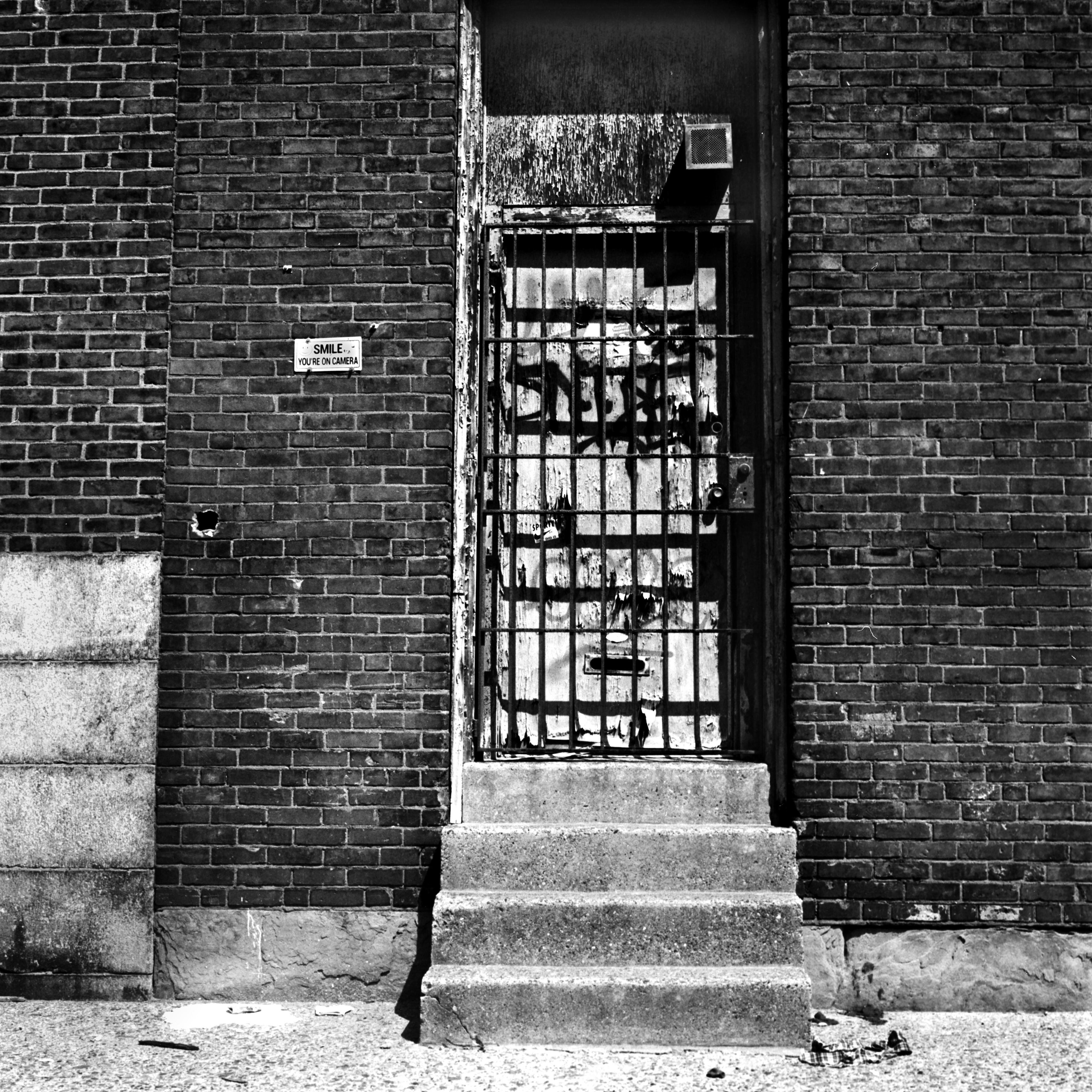 Four steep steps leading to a door with graffiti on it and a mail slit on the bottom. There is a locked gate in front of the door.. On both sides of the door is brick wall with a small sign that reads 'SMILE YOU'RE ON CAMERA'.