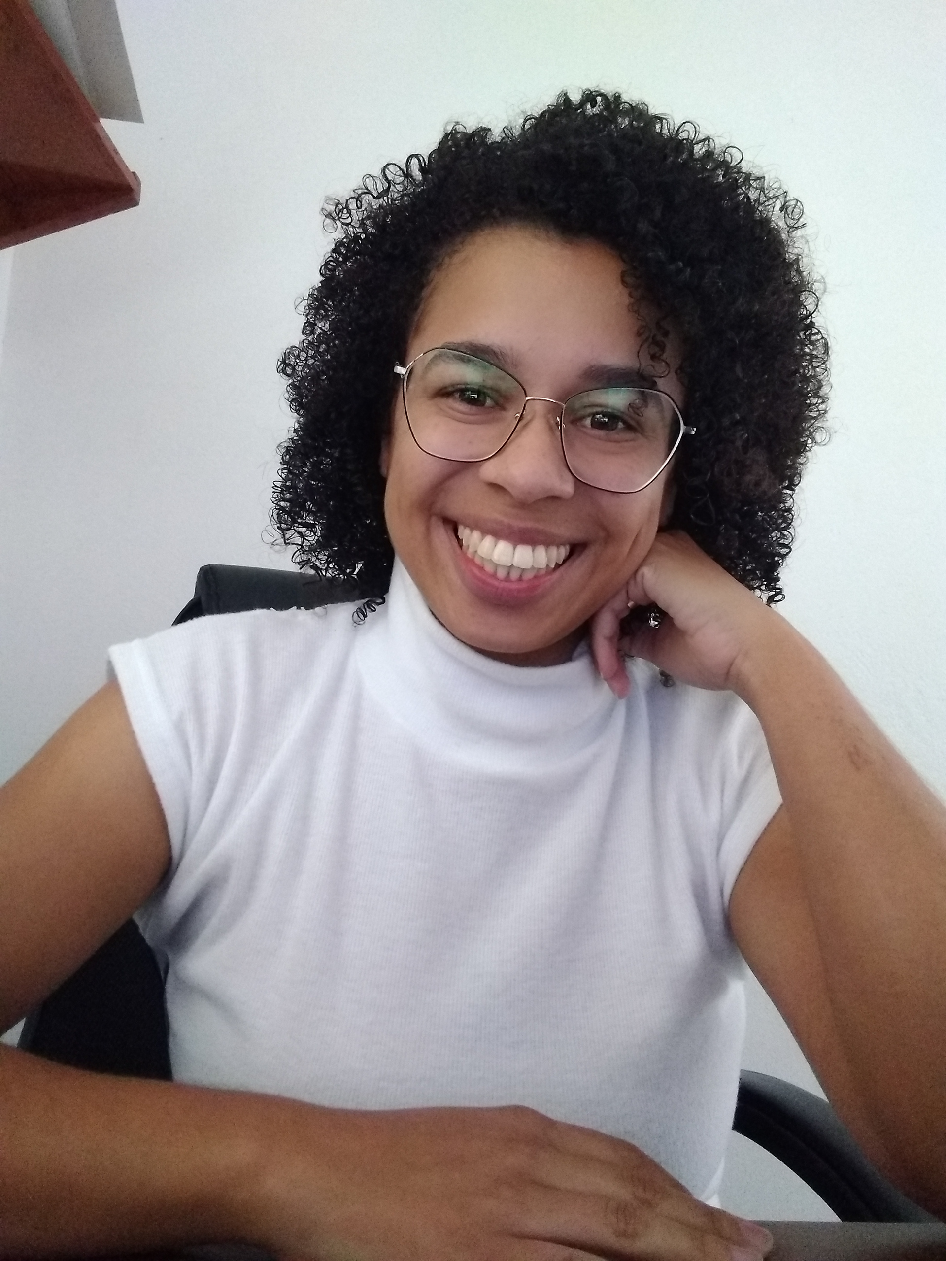 cabelos cacheados até os ombros, óculos de grau, camisa branca com gola alta