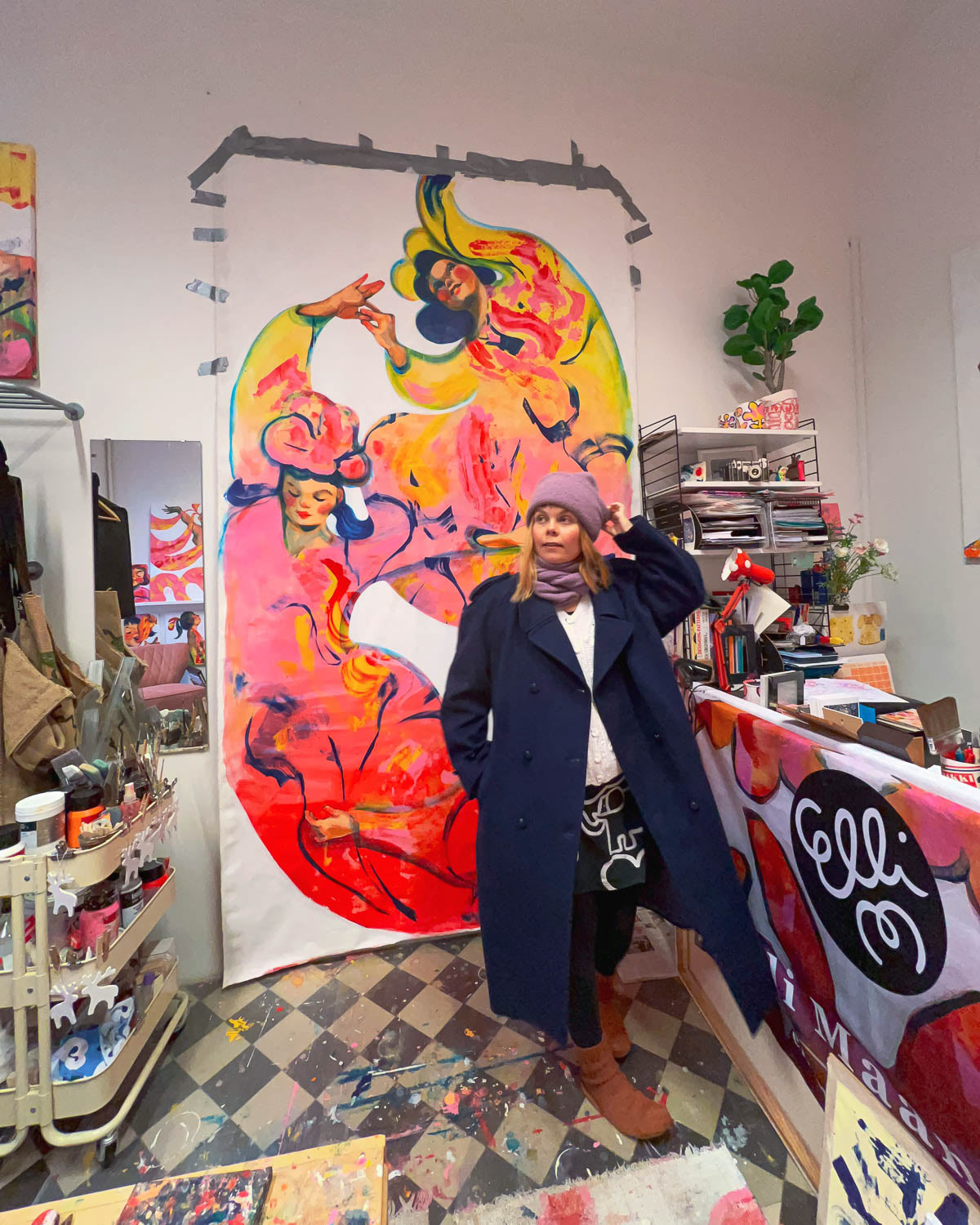 Artist Elli Maanpää in her Helsinki atelier, standing beside her painting The Three Graces, a vibrant large-scale acrylic artwork.
