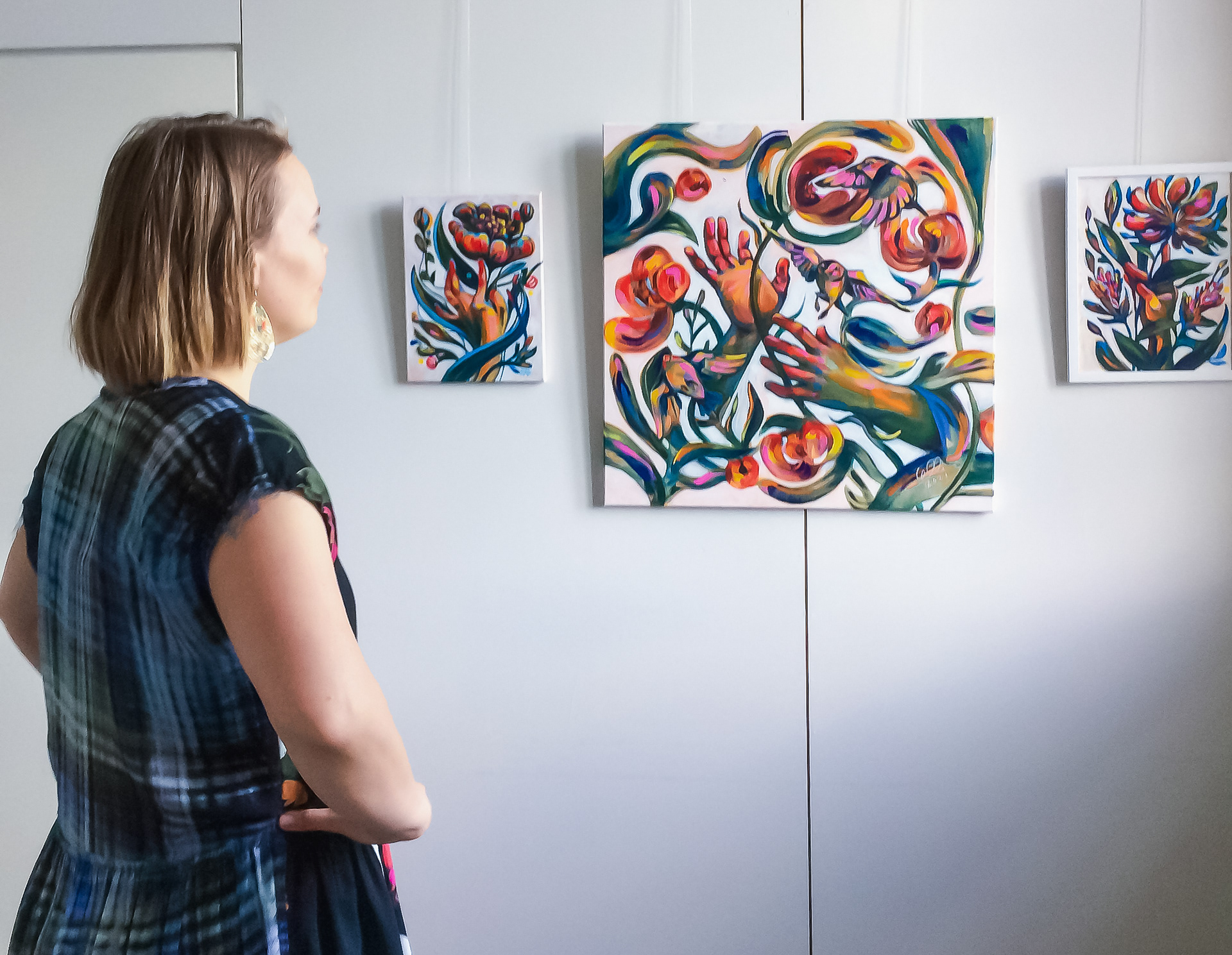 A person standing in a gallery space, thoughtfully looking at colorful paintings by Elli Maanpää on a white wall.