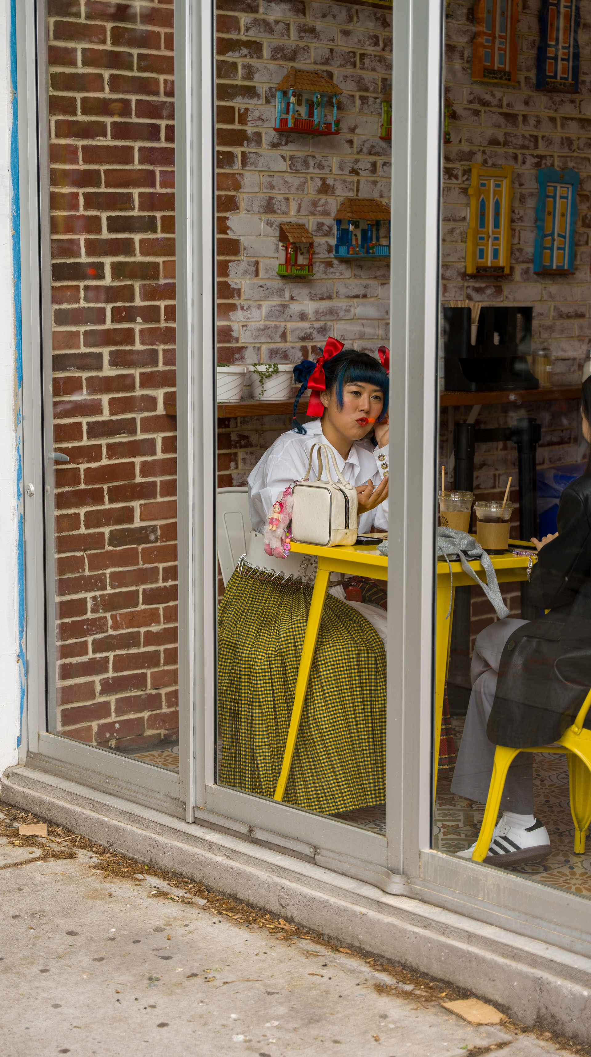 A person with colorful hair and clothing sits at a yellow table inside a café, visible through large glass windows. The café interior features exposed brick walls with bright artwork. is holding a drink and looking toward the window.