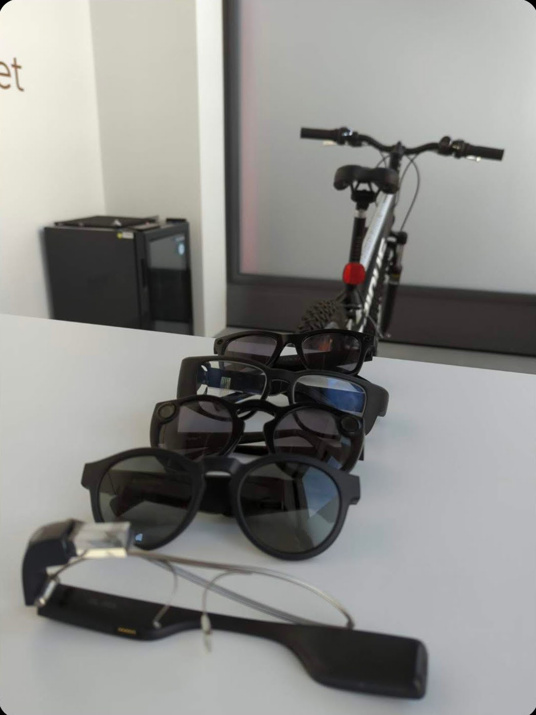 A stack of smart glasses (Google Glass EE1, Bose Frames Rondo, Snap Spectacles 1, Tooz Devkit 20, Ray Ban Stories) laying on a table in front of a bicycle