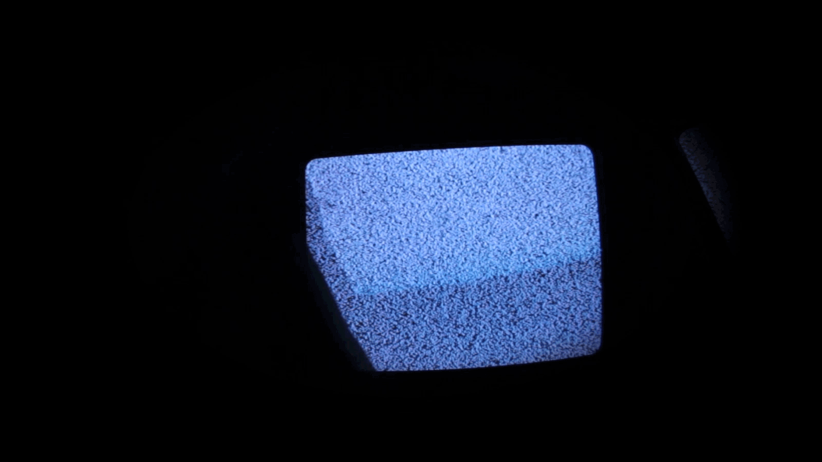 Pequeno vídeo em loop de uma televisão de tubo ao fundo, fora do ar, com chiados. Enquanto isso pessoas passam na frente dela. O ambiente escuro só permite ver a silhueta da pessoa passando.