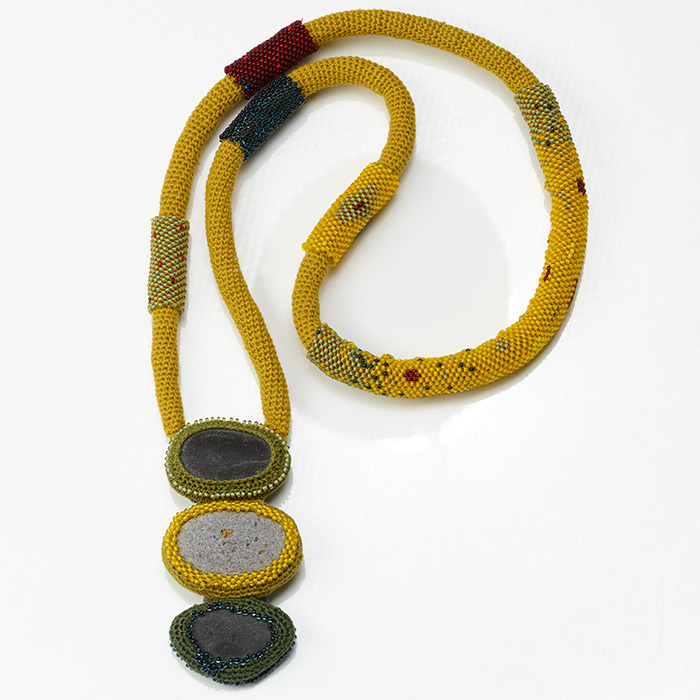 Halsschmuck aus drei Kieselsteinen mit gehäkelter Fassung an langer Kette aus gehäkeltem Schlauch, der teilweise mit Glasperlen umfädelt ist. Die Farben sind Senfgelb, Dunkelrot und verschiedene Grüntöne.