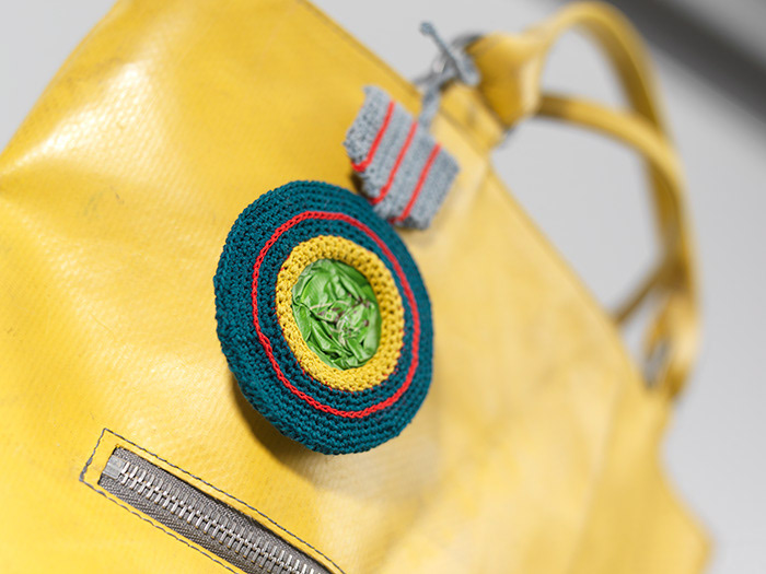 Taschenschmuck, der an einen Orden erinnert, aus flachgehämmerter Kaffeekapsel, mit petrolfarbenem und gelbem Garn in Runden umhäkelt, an der Oberseite graues gehäkeltes Band, und Bindeband
