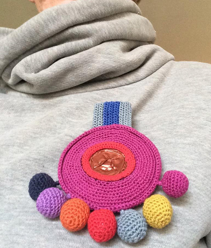 Runde gehäkelte Ordensbrosche in Pink, oben eine Band in Grau und Blau, an der Unterseite baumeln mehrere bunte gehäkelte Kugeln