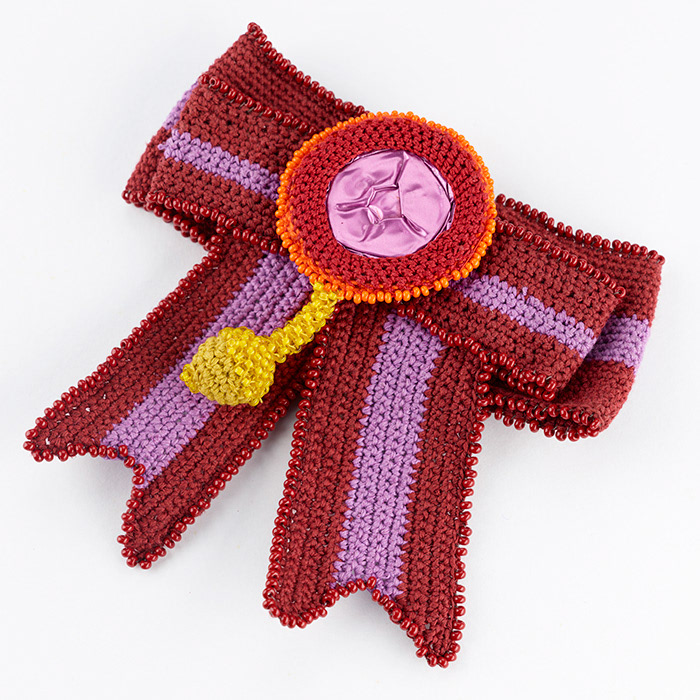 Eine gehäkelte Schleife als Brosche in den Farben Dunkelrot und Helllila. Mittig ist eine runde umhäkelte Kaffeekapsel in Pink platziert, an der eine gelbe gehäkelte Perle hängt, die mit gelben Glasperlen bestickt ist.