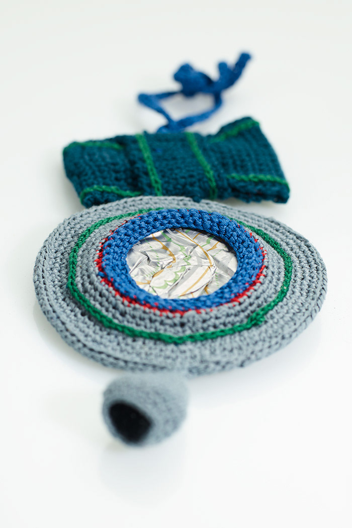 Taschenschmuck aus flachgehämmerter Kaffeekapsel, mit grauem Garn in Runden umhäkelt, an der Oberseite petrolfarbene Schleife, gehäkelt, und Bindeband