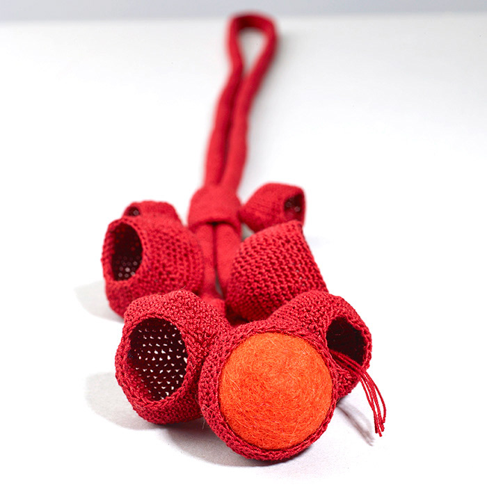 Gehäkelter Halsschmuck in Rot. Mehrere konkave Formen hängen an einem langen gehäkelten Band. Eine Form ist mit einer Filzkugel in Orange gefüllt. Detailbild.