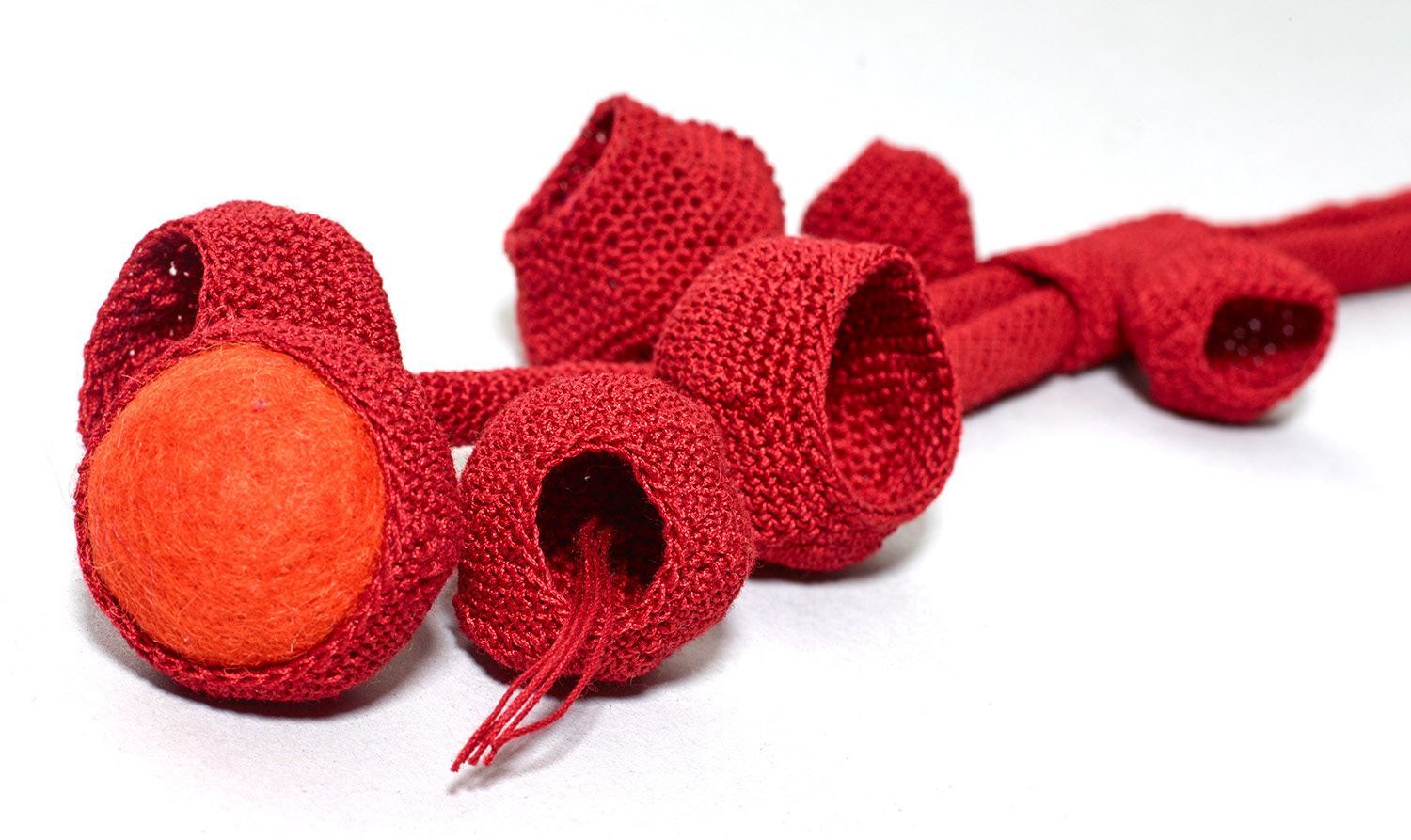 Gehäkelter Halsschmuck in Rot. Mehrere konkave Formen hängen an einem langen gehäkelten Band. Eine Form ist mit einer Filzkugel in Orange gefüllt.