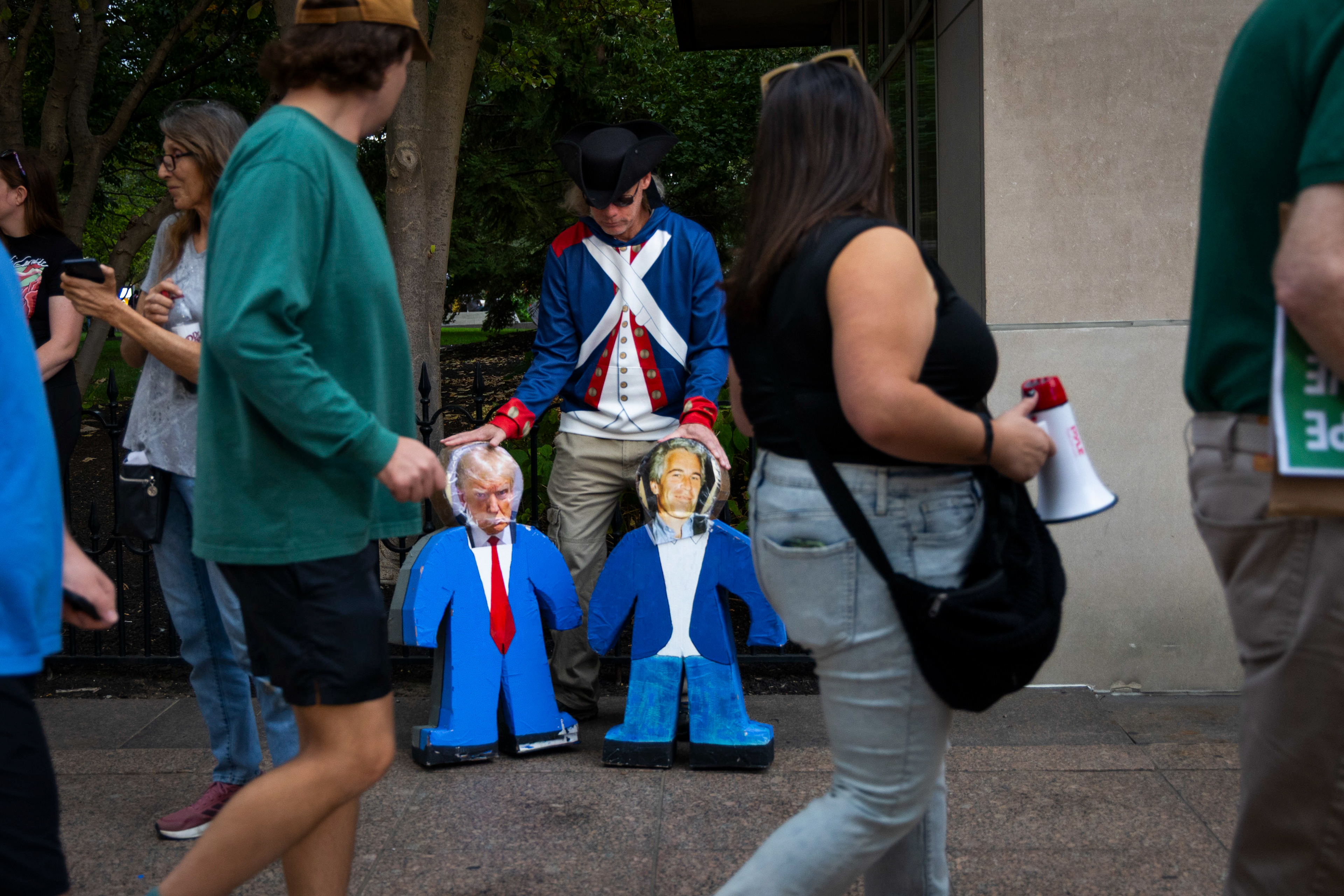 A man displays two human shaped piñatas with photos of Donald Trump and Jeffrey Epstein adhered to them.